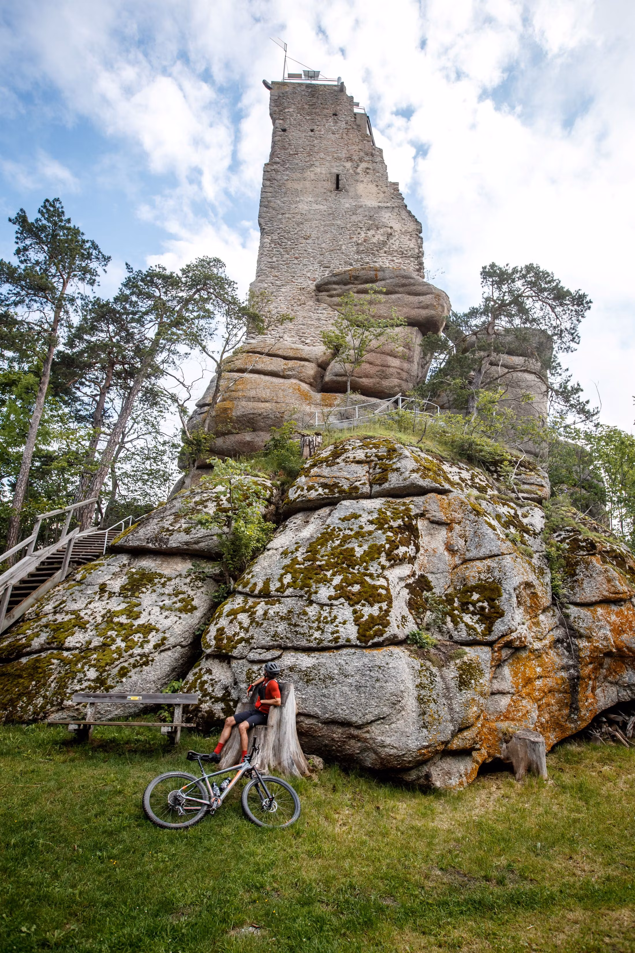 Egy kerékpáros egy facsonknak támaszkodik egy magas, sziklás torony előtt egy erdős területen.