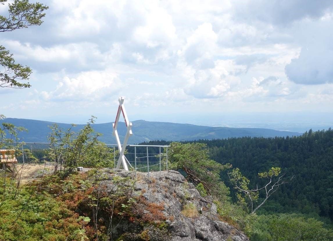 Kilátópont egy hegyre, fehér, kereszt alakú kerettel és felhős égbolt alatti erdős dombokra néző kilátással.