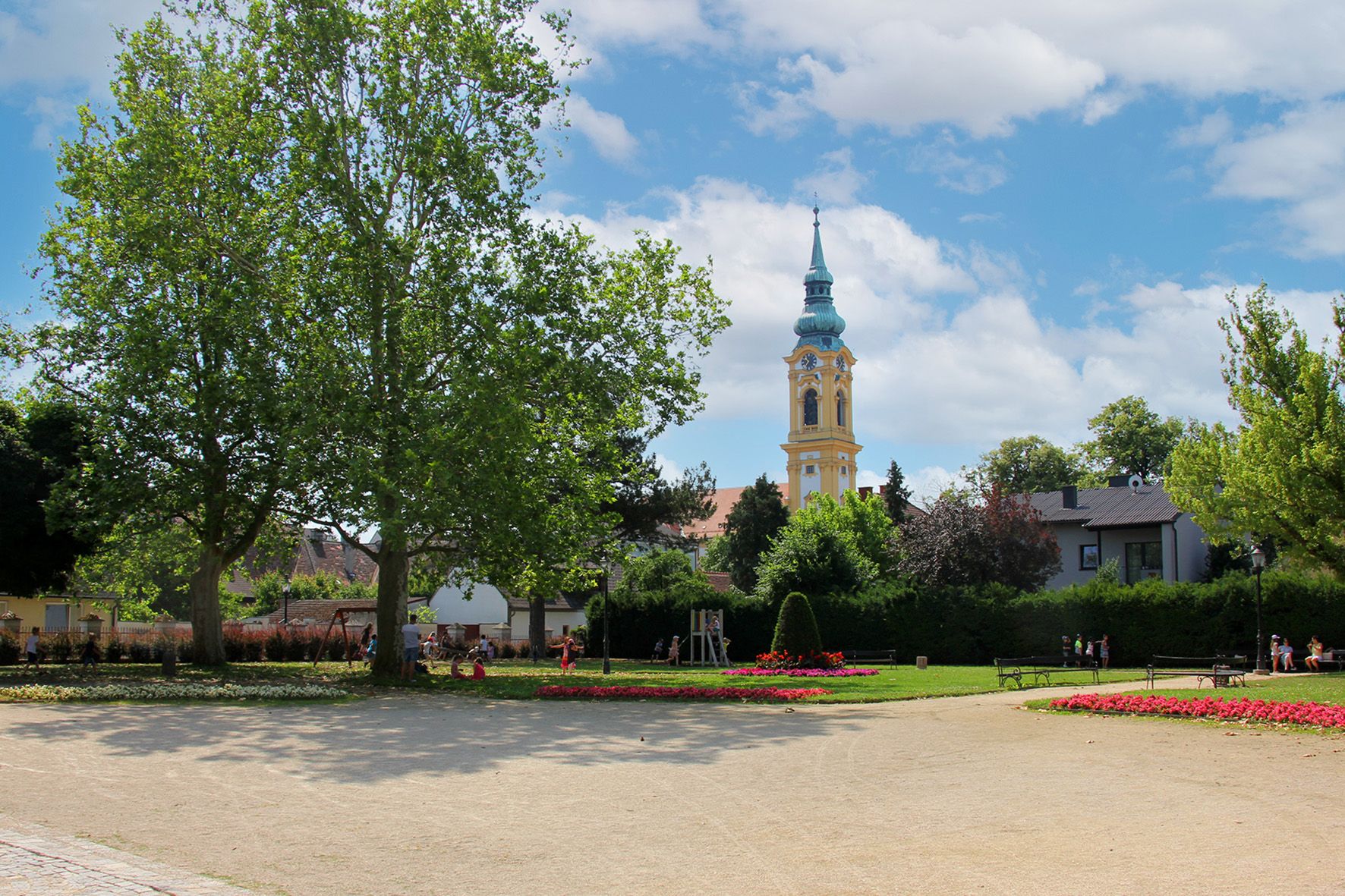 Egy park fákkal, virágágyásokkal és egy templomtoronnyal a háttérben.