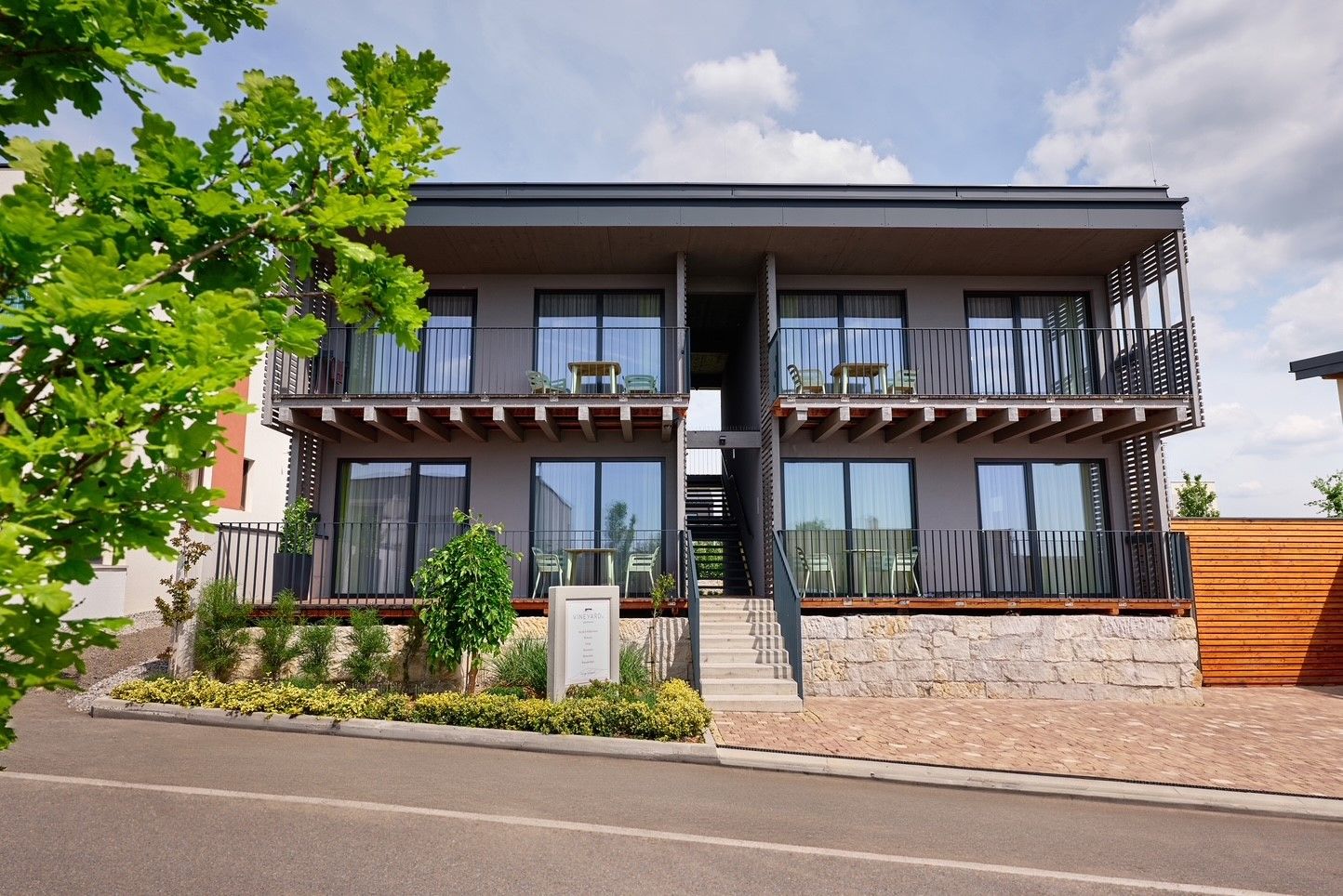 Modern, kétszintes, erkélyes, lépcsőházzal ellátott, növényekkel és fákkal körülvett, kétszintes apartmanegyüttes.