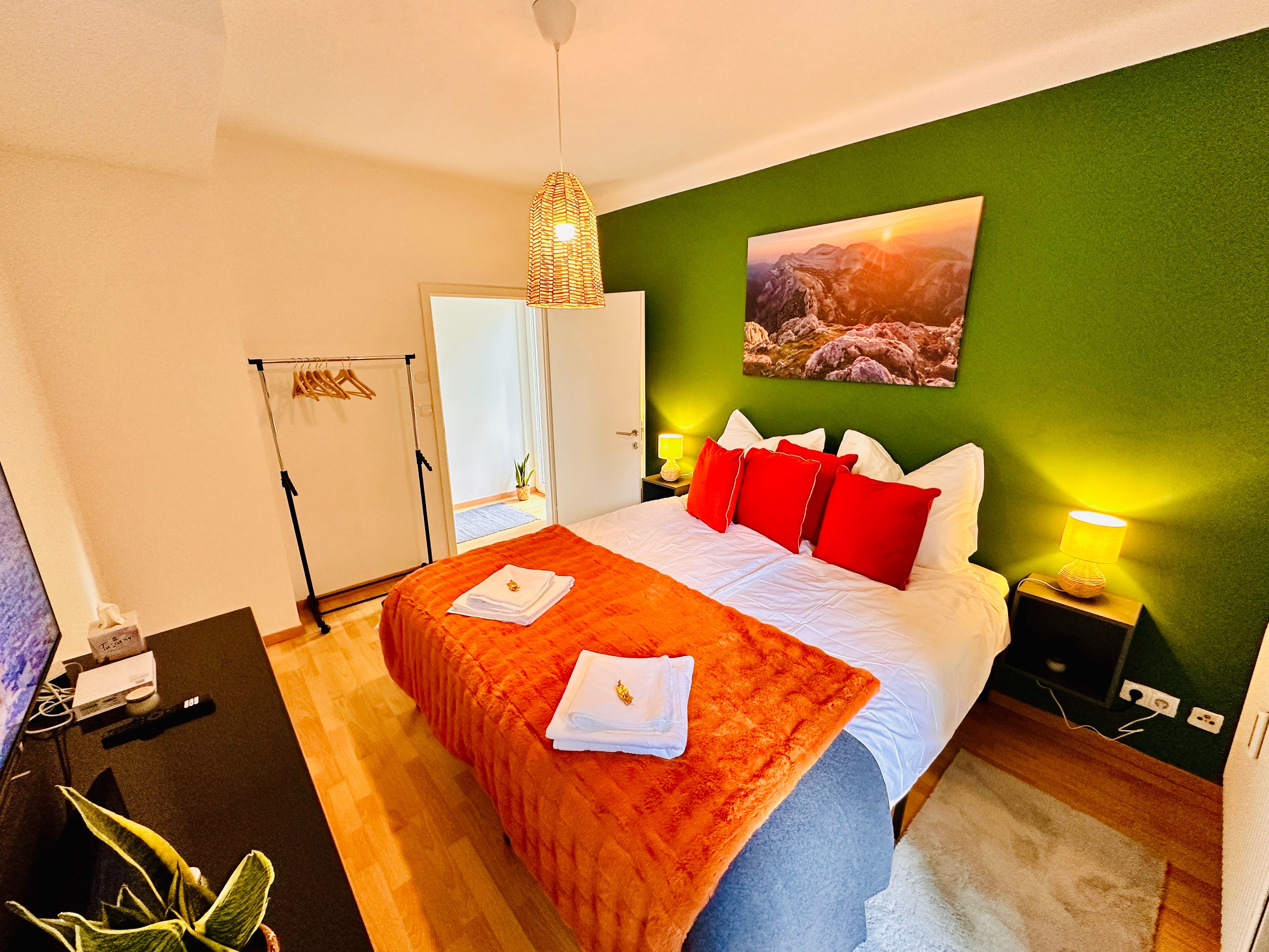 Hangulatos hálószoba zöldre festett fallal, franciaággyal, piros párnákkal, éjjeliszekrényekkel és lámpával.