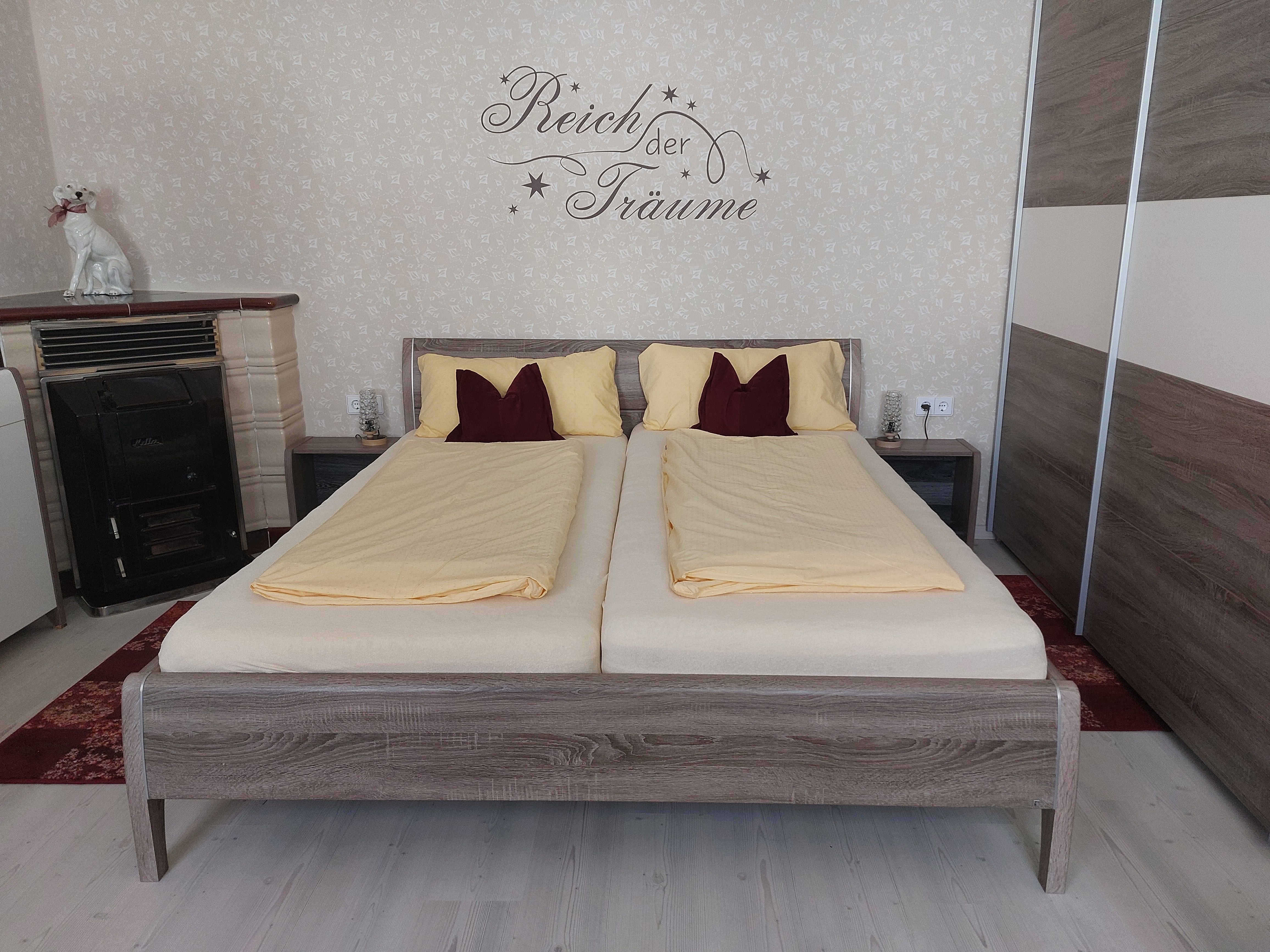 Egy hálószoba franciaággyal, sárga ágyneművel és írással a falon. Egy kályha és egy kutyaszobor is látható.