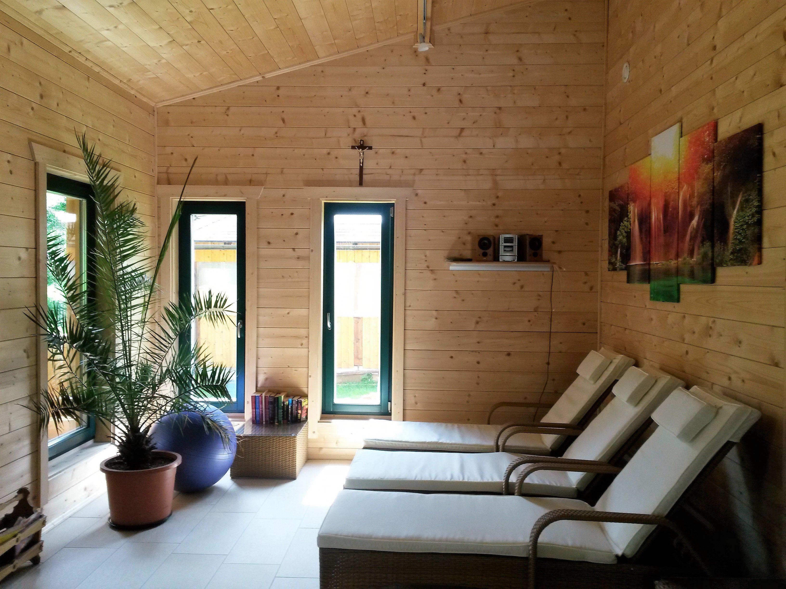 Egy szauna belseje, fa burkolattal, nyugágyakkal, pálmafával és egy képpel a falon.