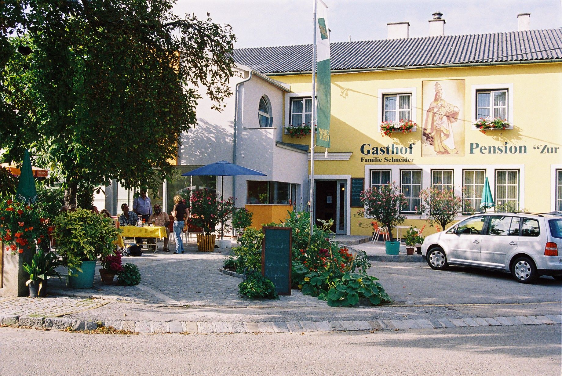 A "Gasthof-Pension Zur Hammerschmiede" feliratú sárga épület. Az épület előtt asztalok, székek és egy autó áll.
