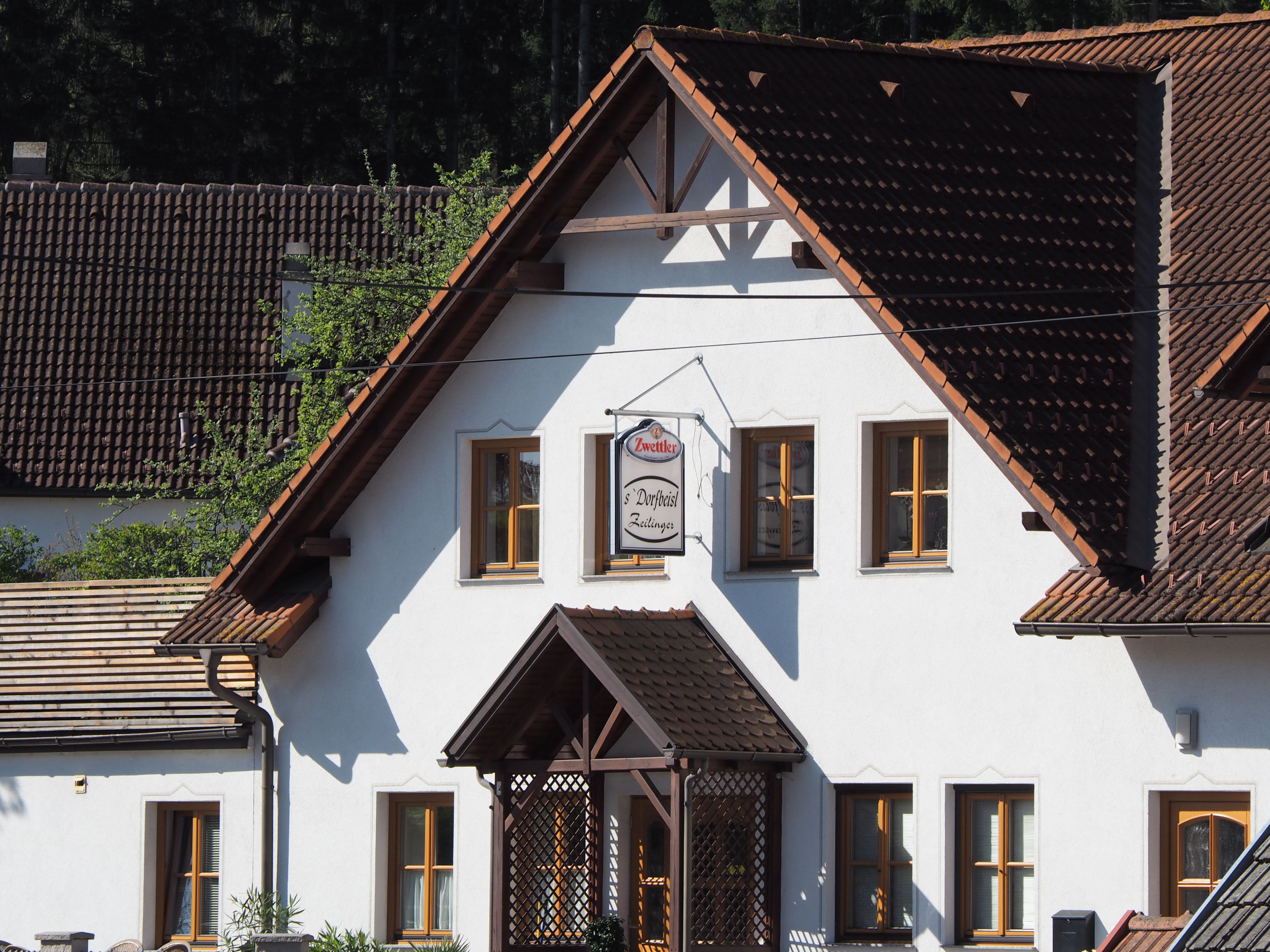 Hagyományos épület fehér falakkal és barna tetőcseréppel. A homlokzaton "Zwettler Dorfwirt Zeilinger" feliratú tábla lóg.