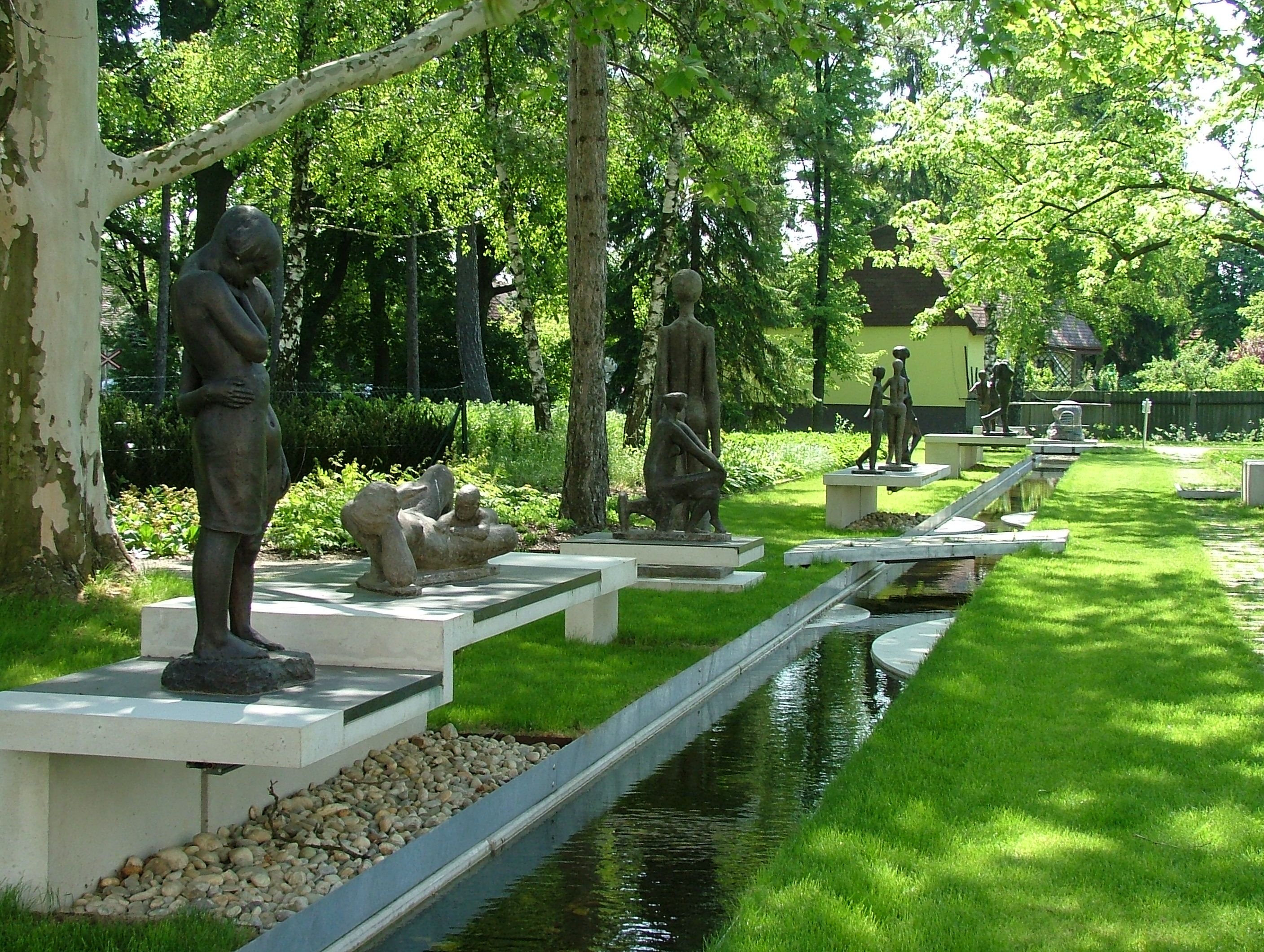 Szobrok Bad Pirawarth parkjában, fákkal és egy kis patakkal körülvéve.