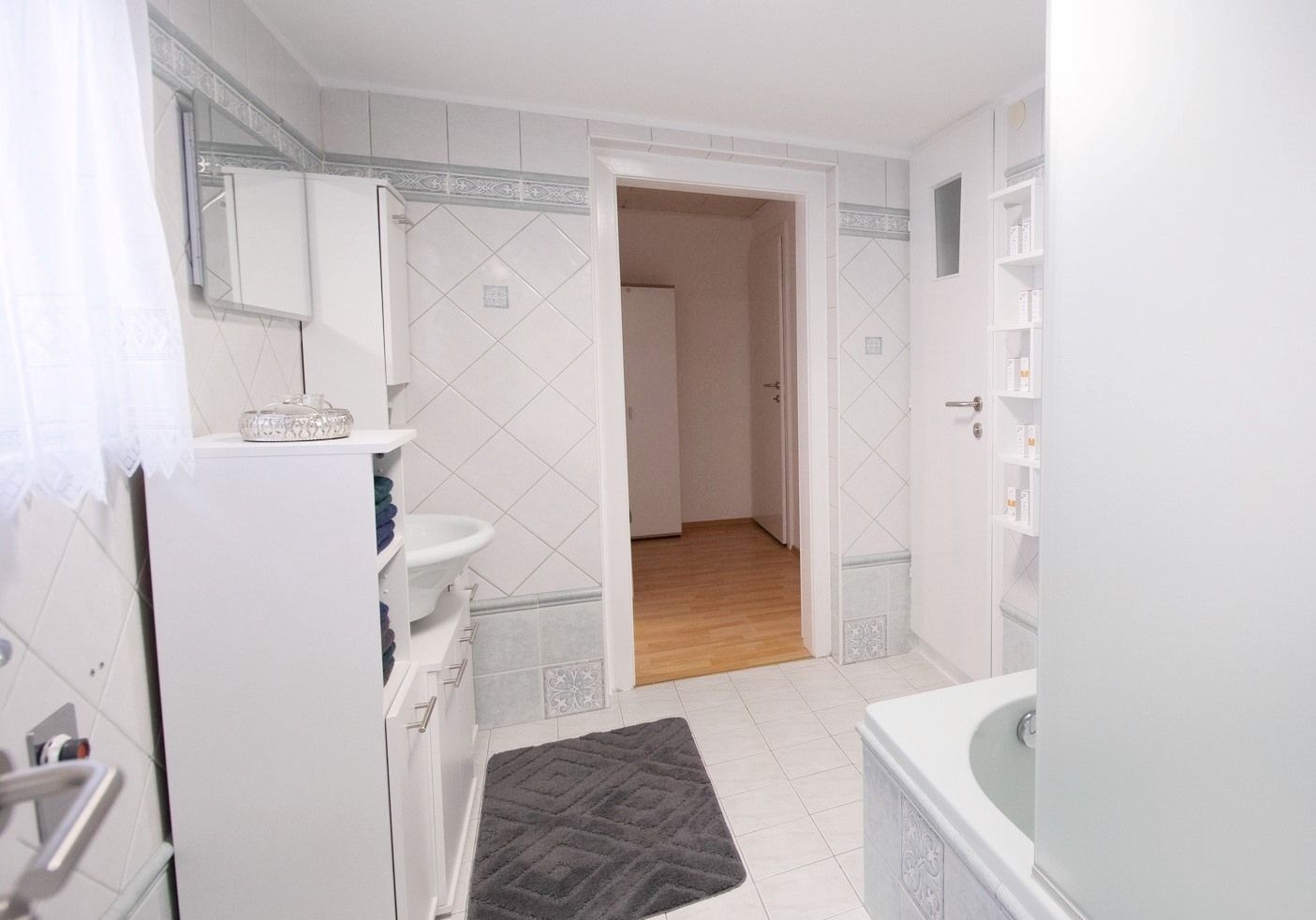 Modern fürdőszoba fehér csempével, káddal, mosdókagylóval és szekrénnyel. Ajtó vezet a fapadlós előszobába.