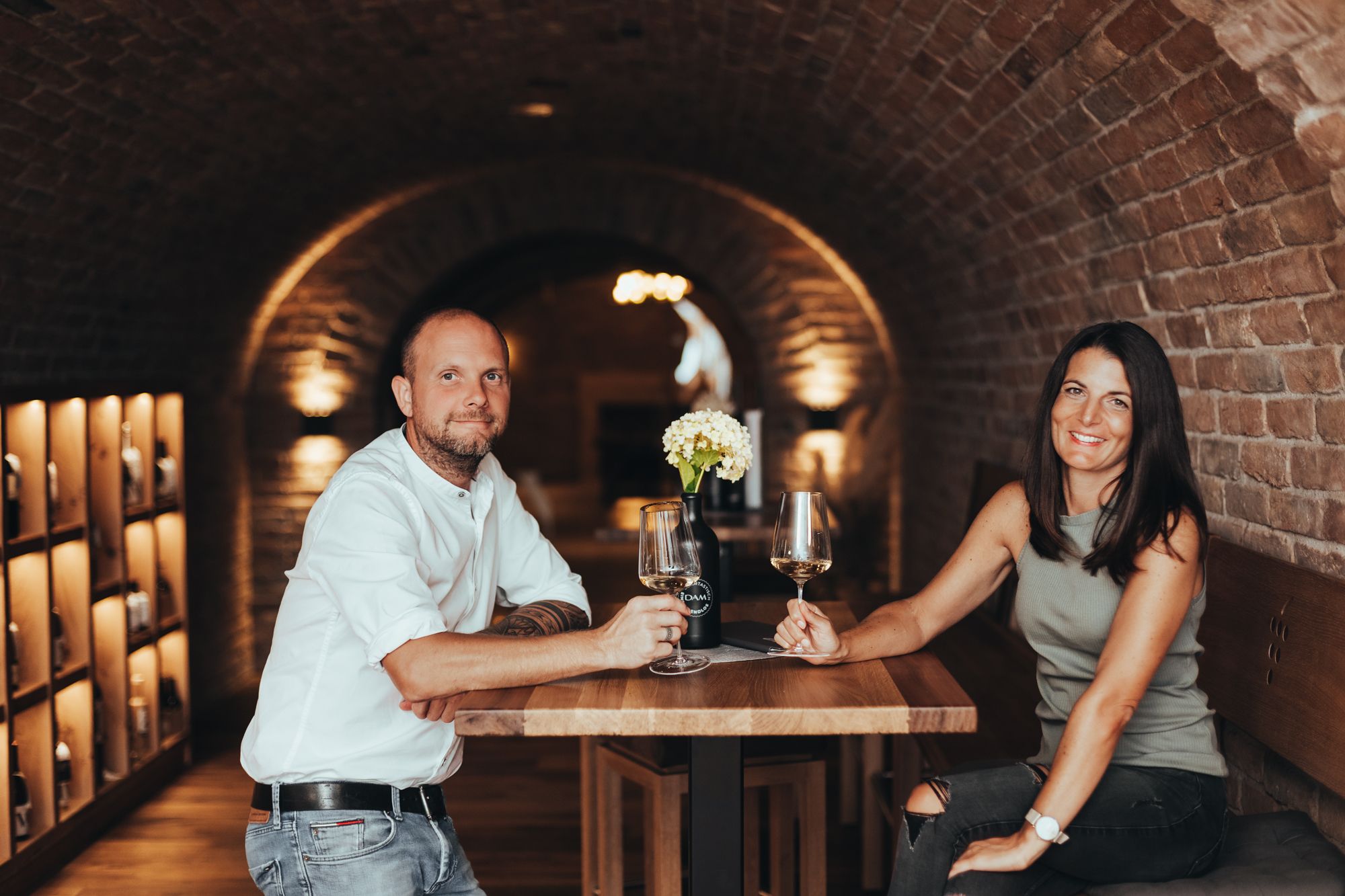 Egy férfi és egy nő egy borospincében egy asztalnál ül, kezükben pohár borral.