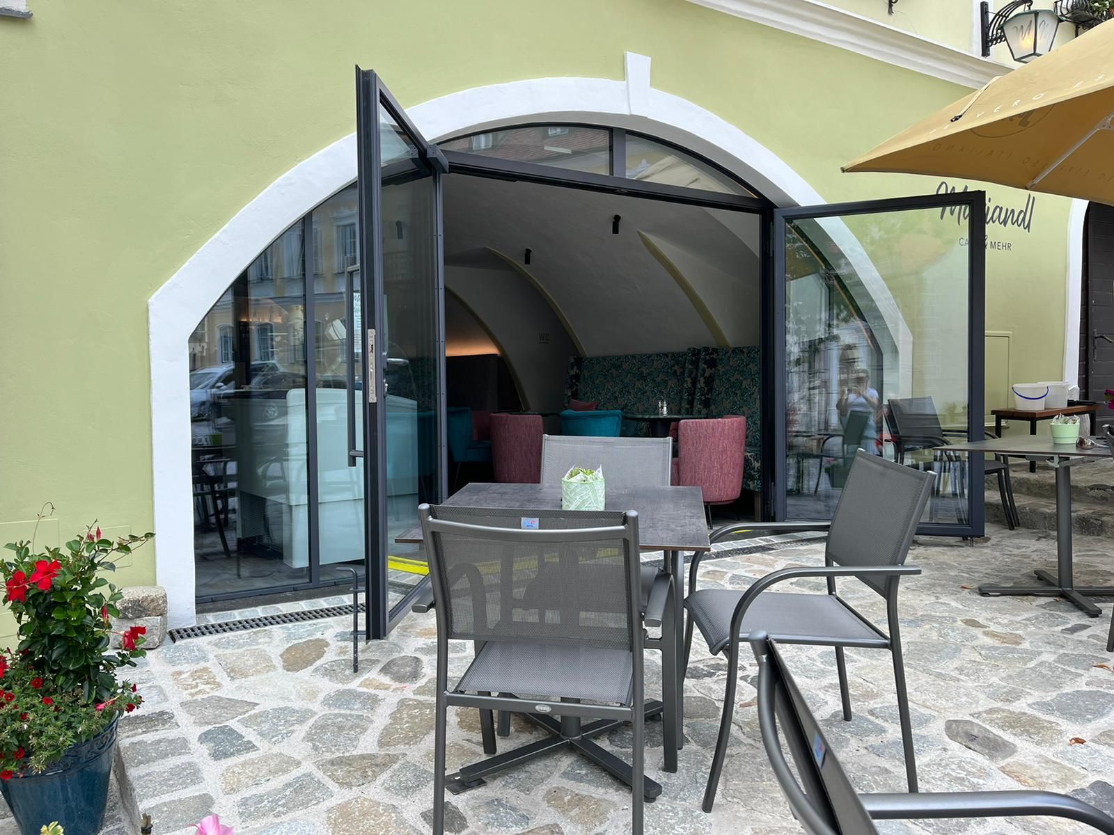Egy kávézó külső nézete nyitott üvegajtókkal és szabadtéri ülőhelyekkel.