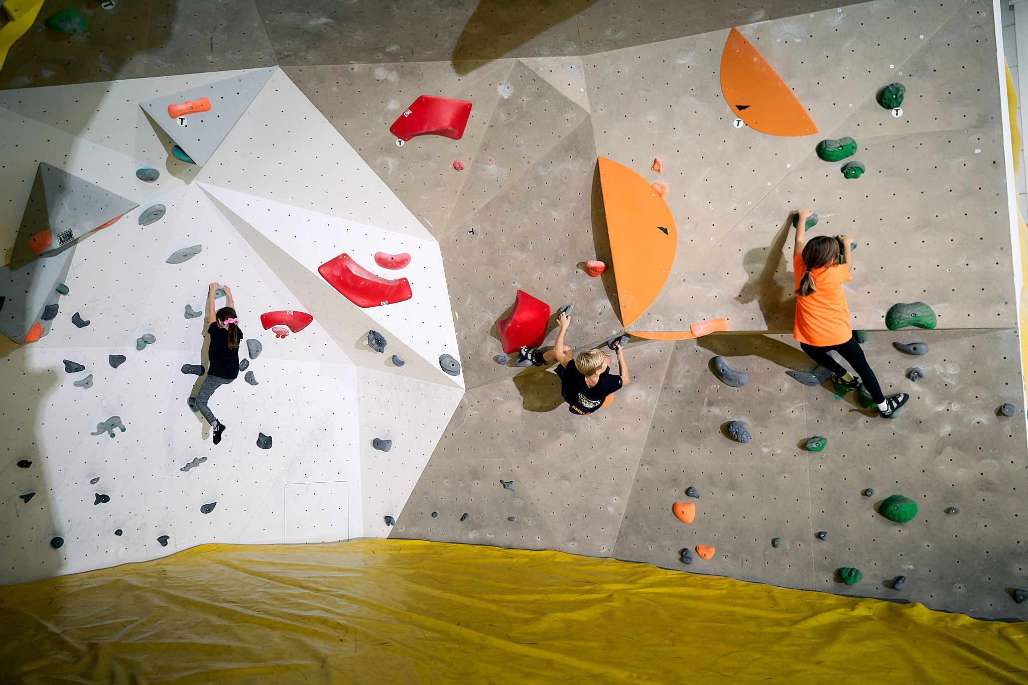 Három gyerek mászik egy beltéri boulderfalon, színes fogásokkal.