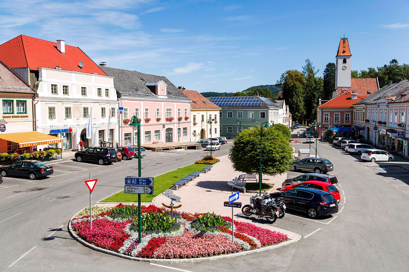 Az Aspang Markt főtere színes virágágyásokkal, parkoló autókkal és történelmi épületekkel.