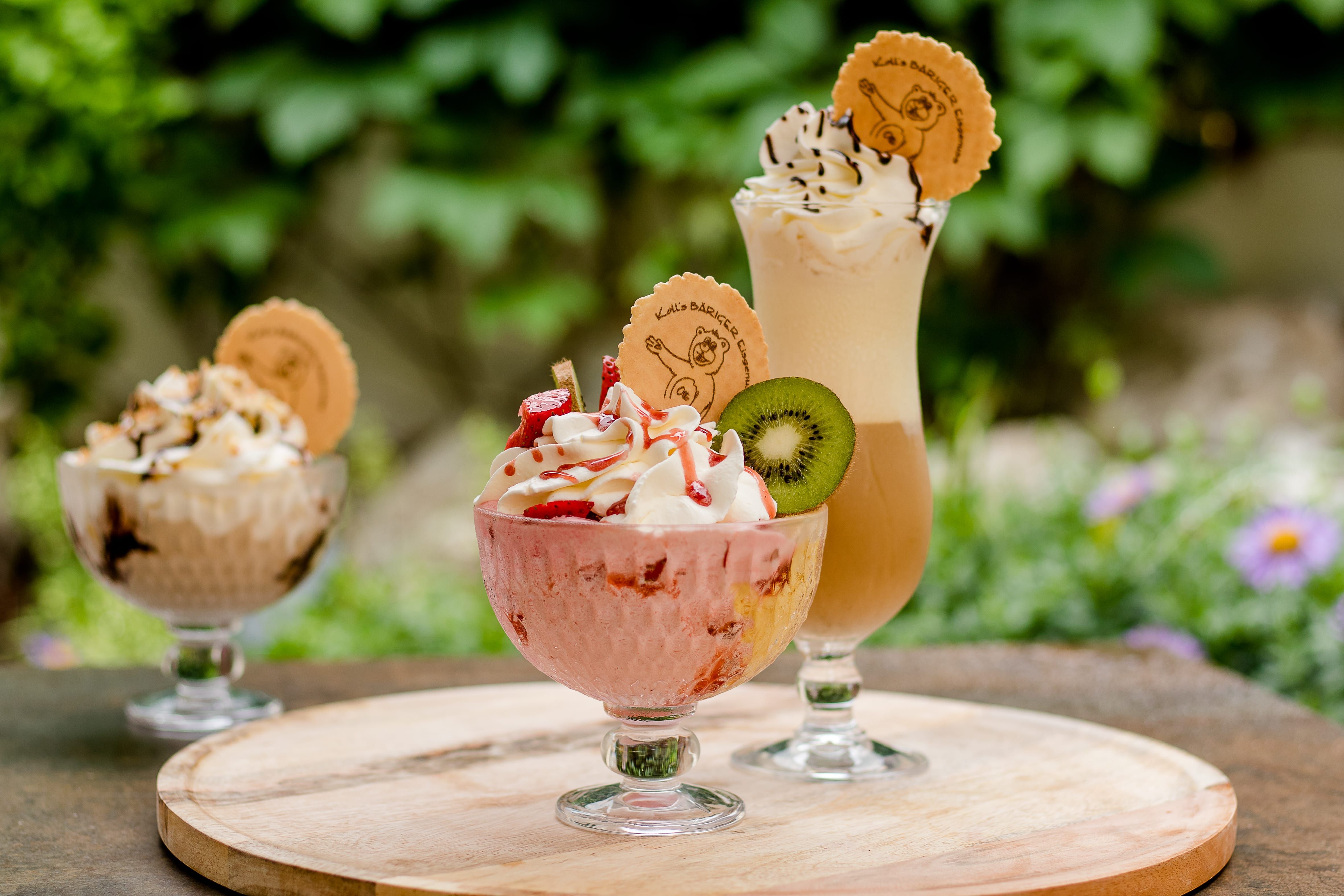 Három fagylaltkehely krémmel és díszítéssel egy faasztalon odakint.