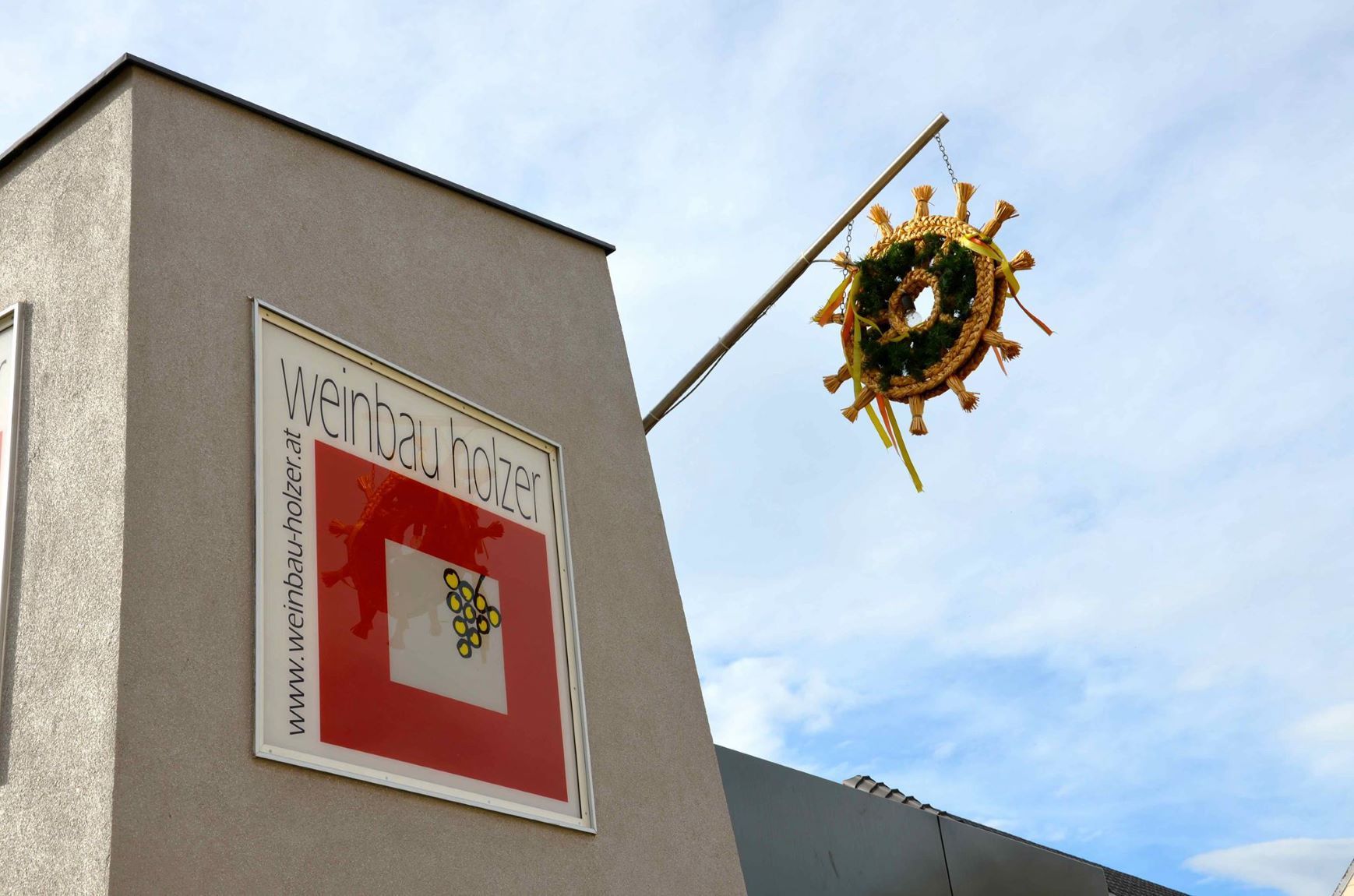 Épület "Weinbau Holzer" felirattal és díszes koszorúval egy oszlopon.
