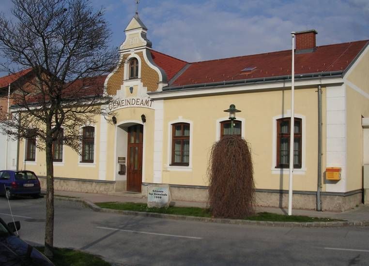 Sárga épület "Gemeindeamt" felirattal, piros tetővel, előtte fa és lámpa.