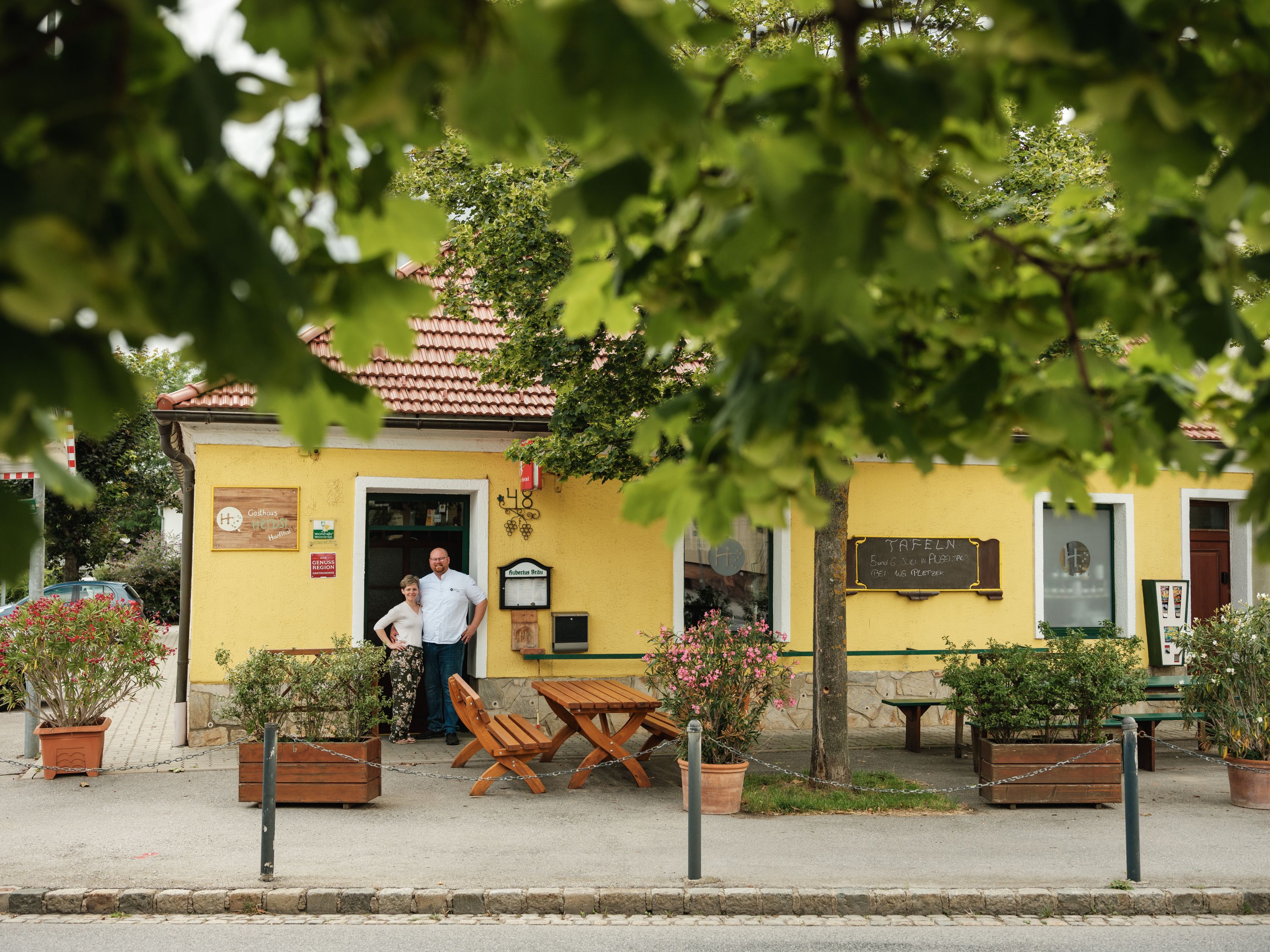 Egy sárga, piros tetejű fogadó, fákkal körülvéve. Két ember áll a bejárat előtt.