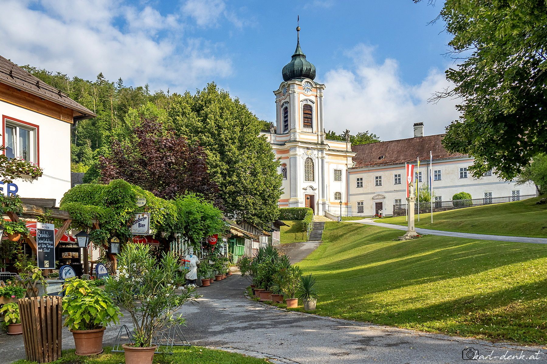 Egy festői falu, előtérben egy barokk stílusú templommal és egy vendéglővel, zöld fák és kék ég alatt.