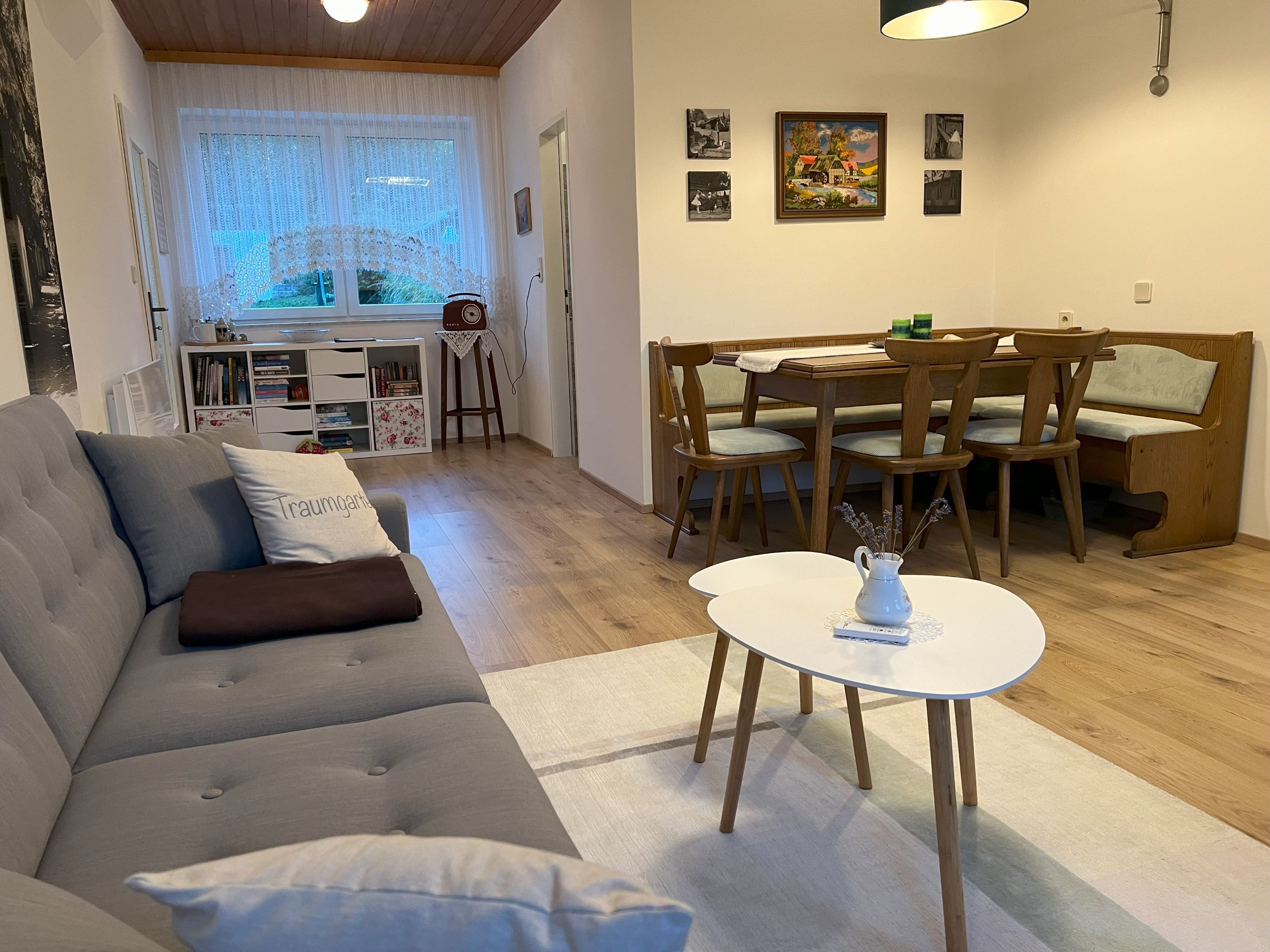 Hangulatos nappali szürke kanapéval, étkezőasztallal és székekkel, könyvespolccal és függönyös ablakkal.