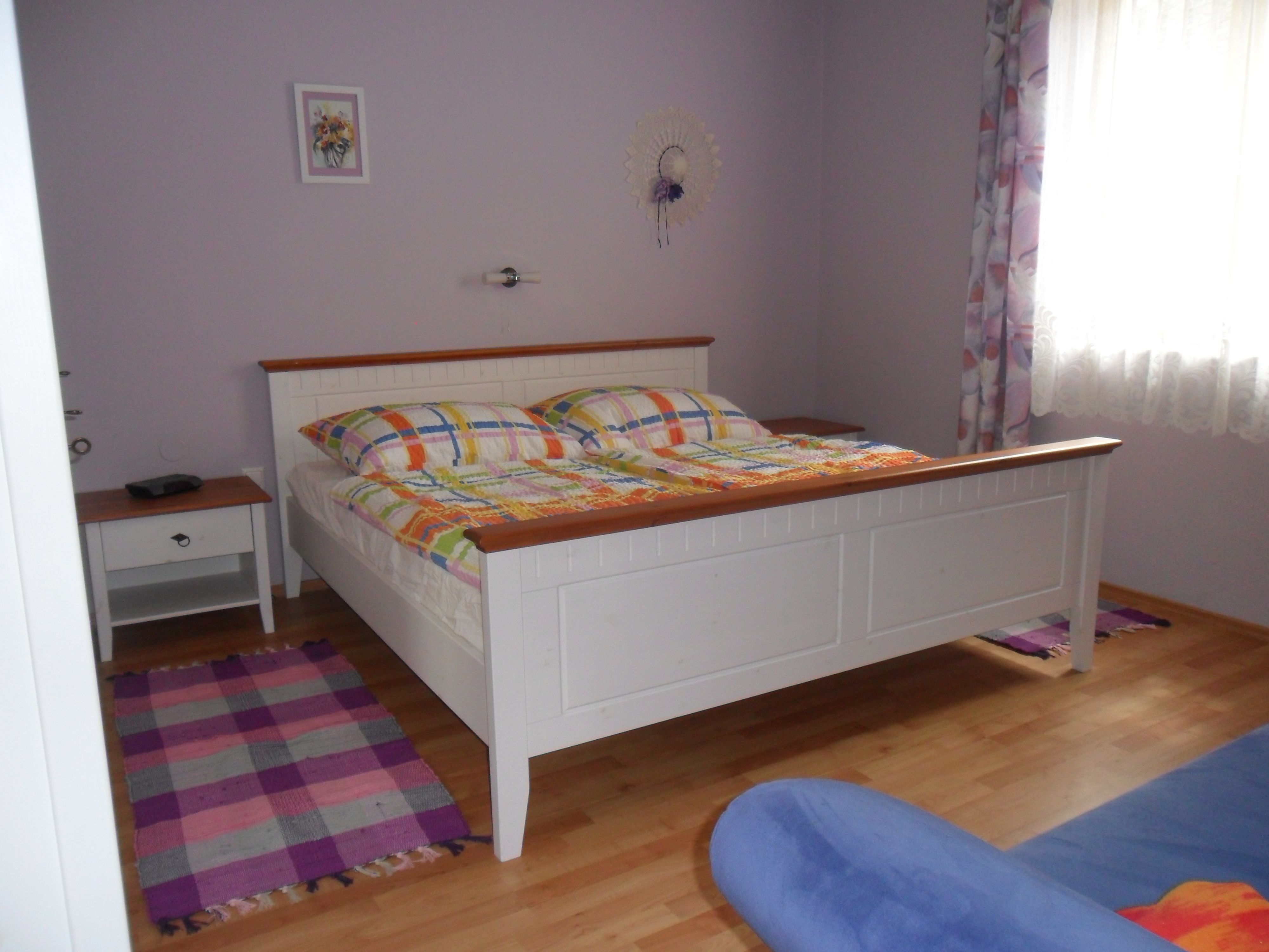 Egy hálószoba fehér ággyal, színes paplanokkal, éjjeliszekrénnyel és függönyös ablakkal.