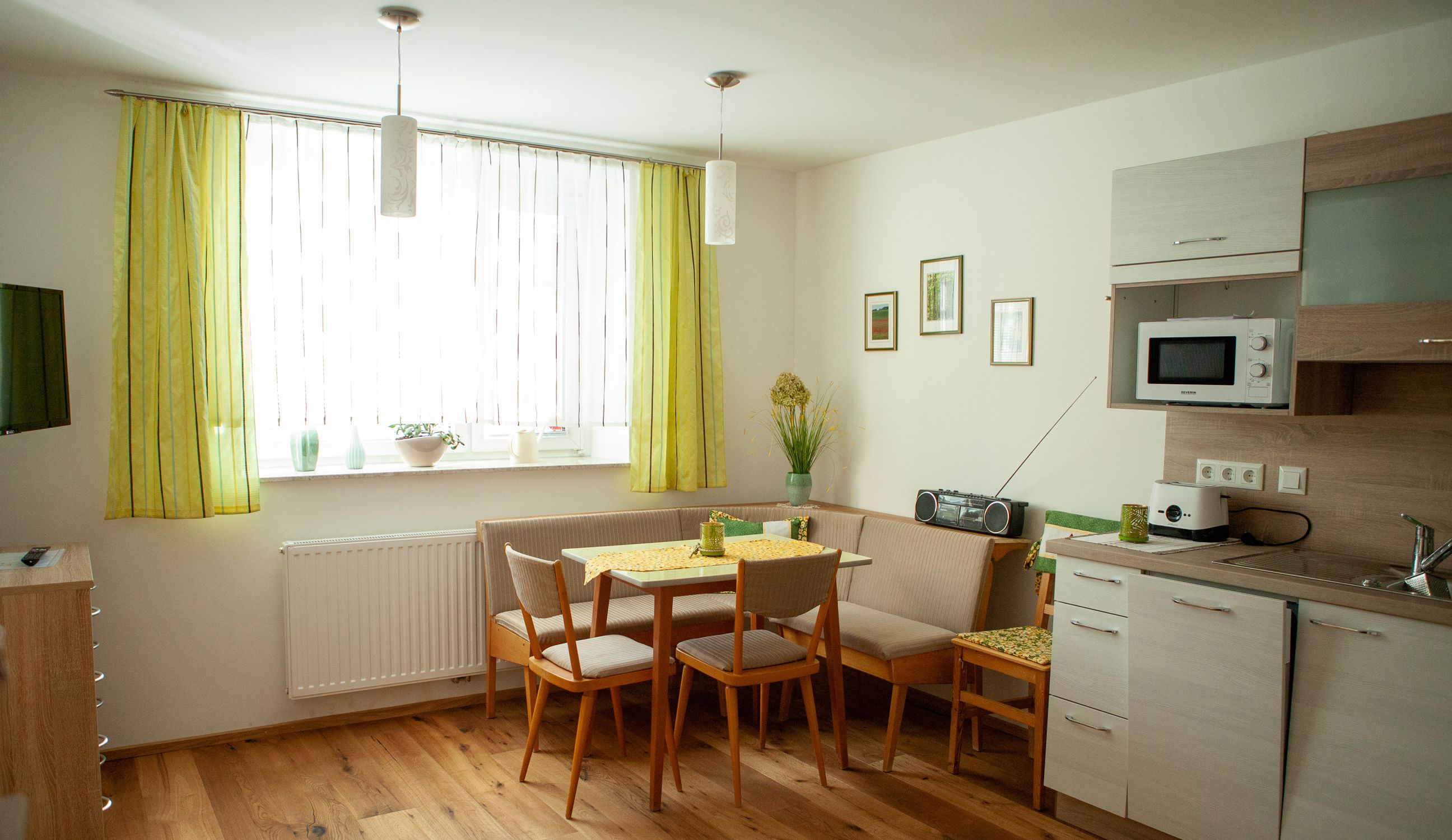 Hangulatos étkezős konyha étkezőasztallal, sarokpaddal, konyhabútorral és zöld függönyökkel.