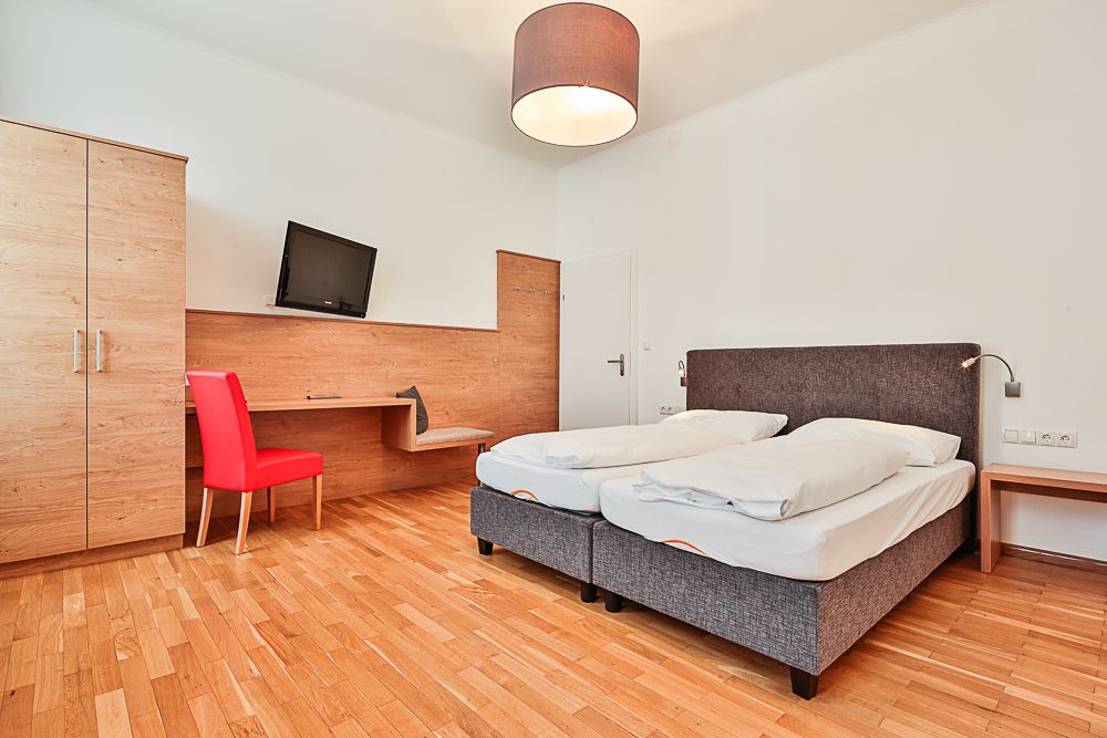 Hotelszoba franciaággyal, piros székkel, íróasztallal és televízióval.