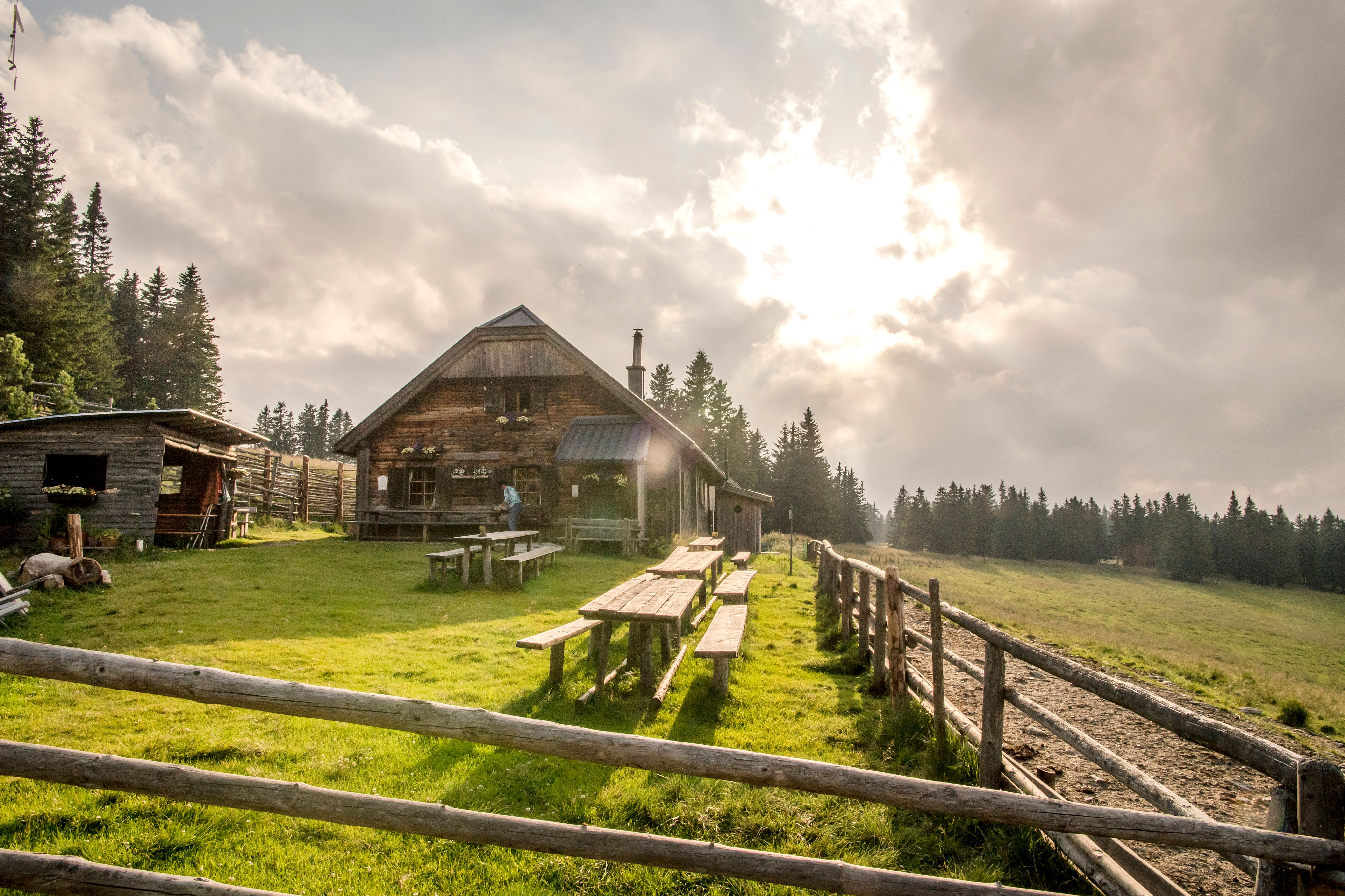 Alpesi hütte faasztalokkal egy réten, erdővel körülvéve, felhős égbolt alatt.