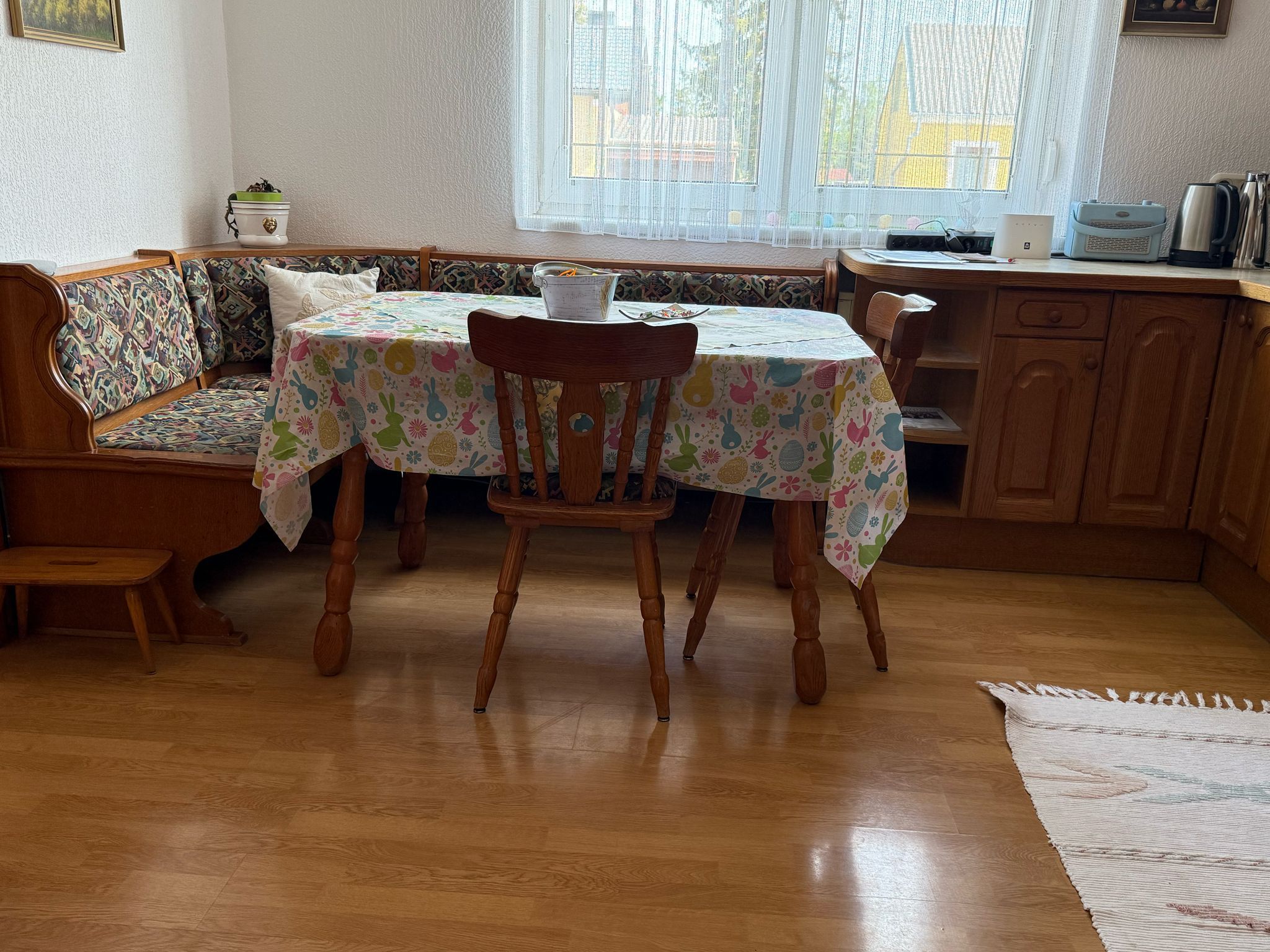 Étkezőhelyiség fa asztallal, székekkel és sarokpaddal, színes terítővel, a háttérben ablak.