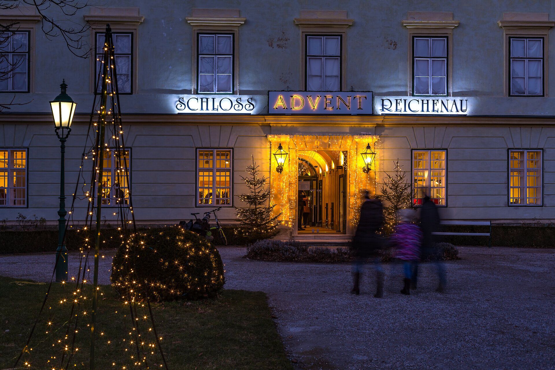 Adventmarkt im Schloss Reichenau an der Rax. Das Schloss ist von außen stimmungsvoll beleuchtet, im Innenhof stehen die Hütten. Menschen stehen davor und trinken Punsch.