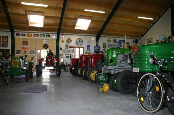 Egy pajta belső nézete, több különböző színű vintage traktorral, a falakon retro reklámtáblákkal körülvéve.