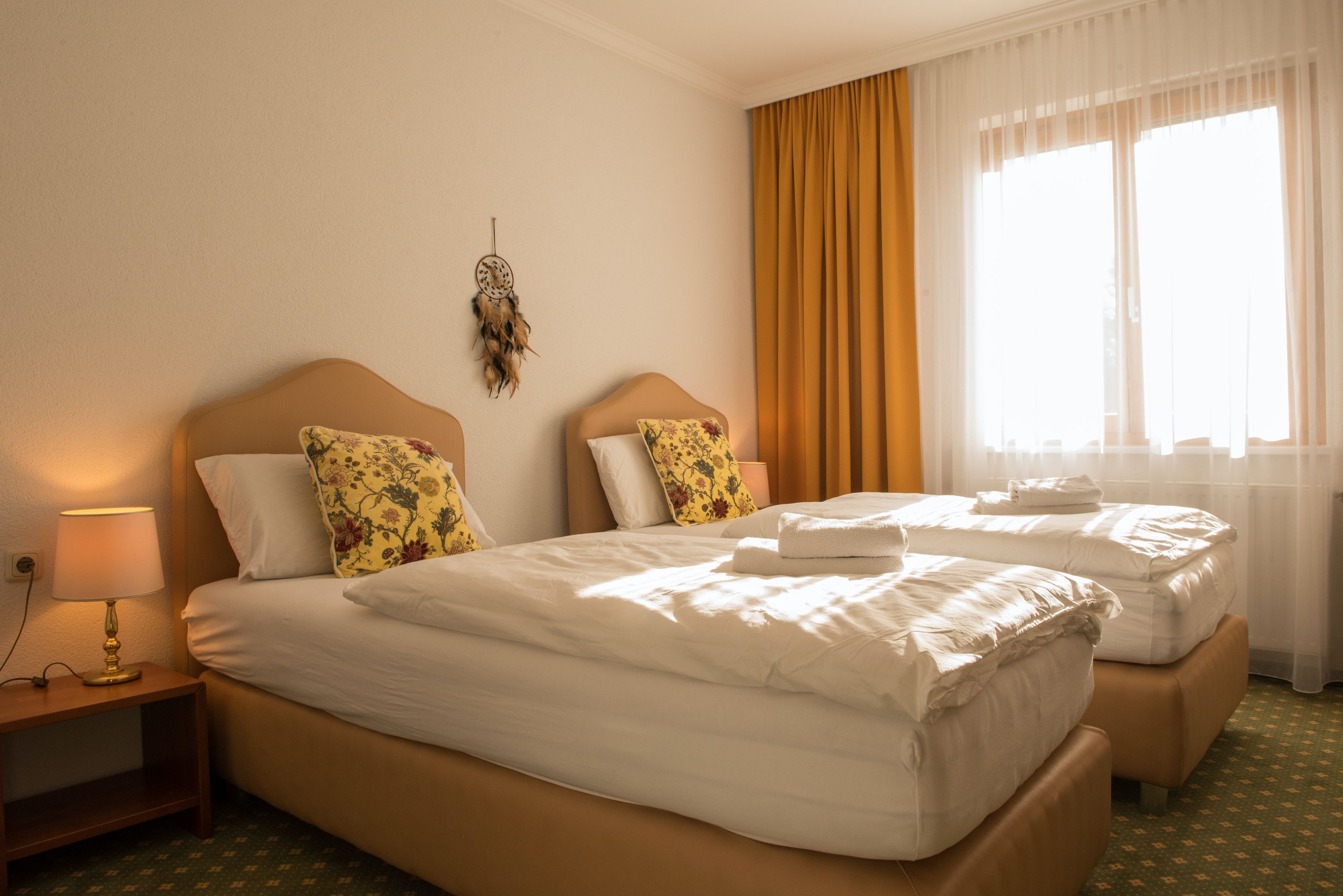 Hangulatos kétágyas szoba két egyszemélyes ággyal, sárga függönyökkel és egy álomfogóval a falon.