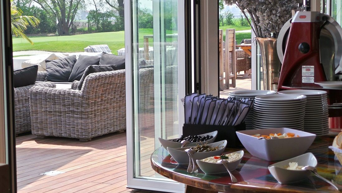 Egy terített asztal, edényekkel és harapnivalókkal, egy rattan bútorokkal berendezett terasz mellett, kilátással a zöld gyepre.
