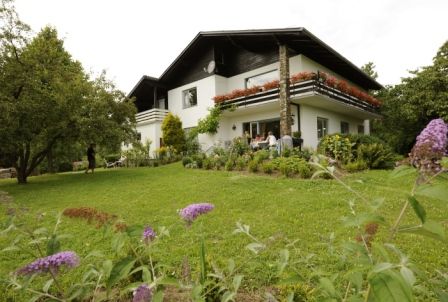 Egy kétszintes, fehér ház erkéllyel, amelyet egy zöld kert vesz körül virágokkal és fákkal.