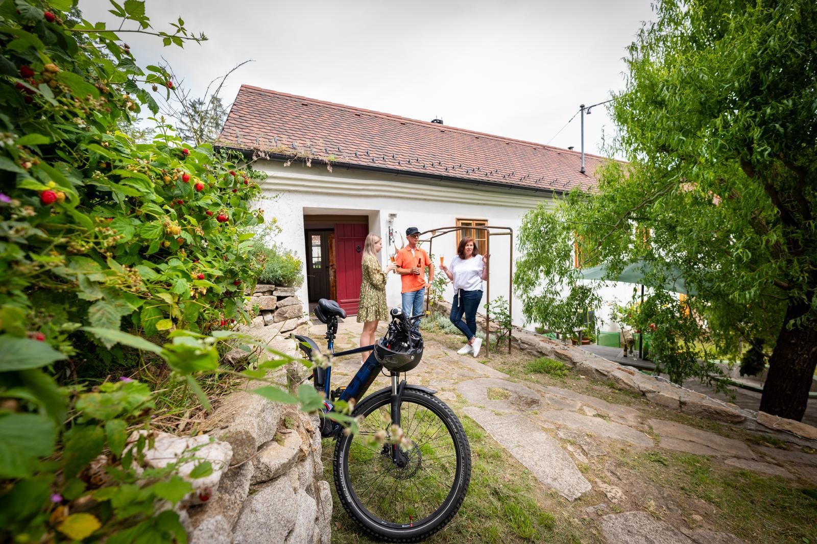 Három ember áll egy fehér, piros tetejű ház előtt, az előtérben egy kerékpár.