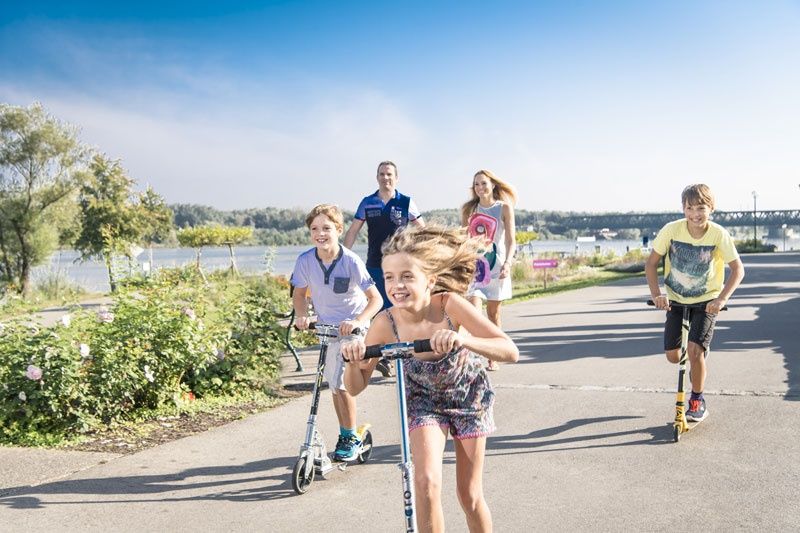 Gyerekek robogóznak egy folyó menti ösvényen, a háttérben két felnőtt kíséretében.