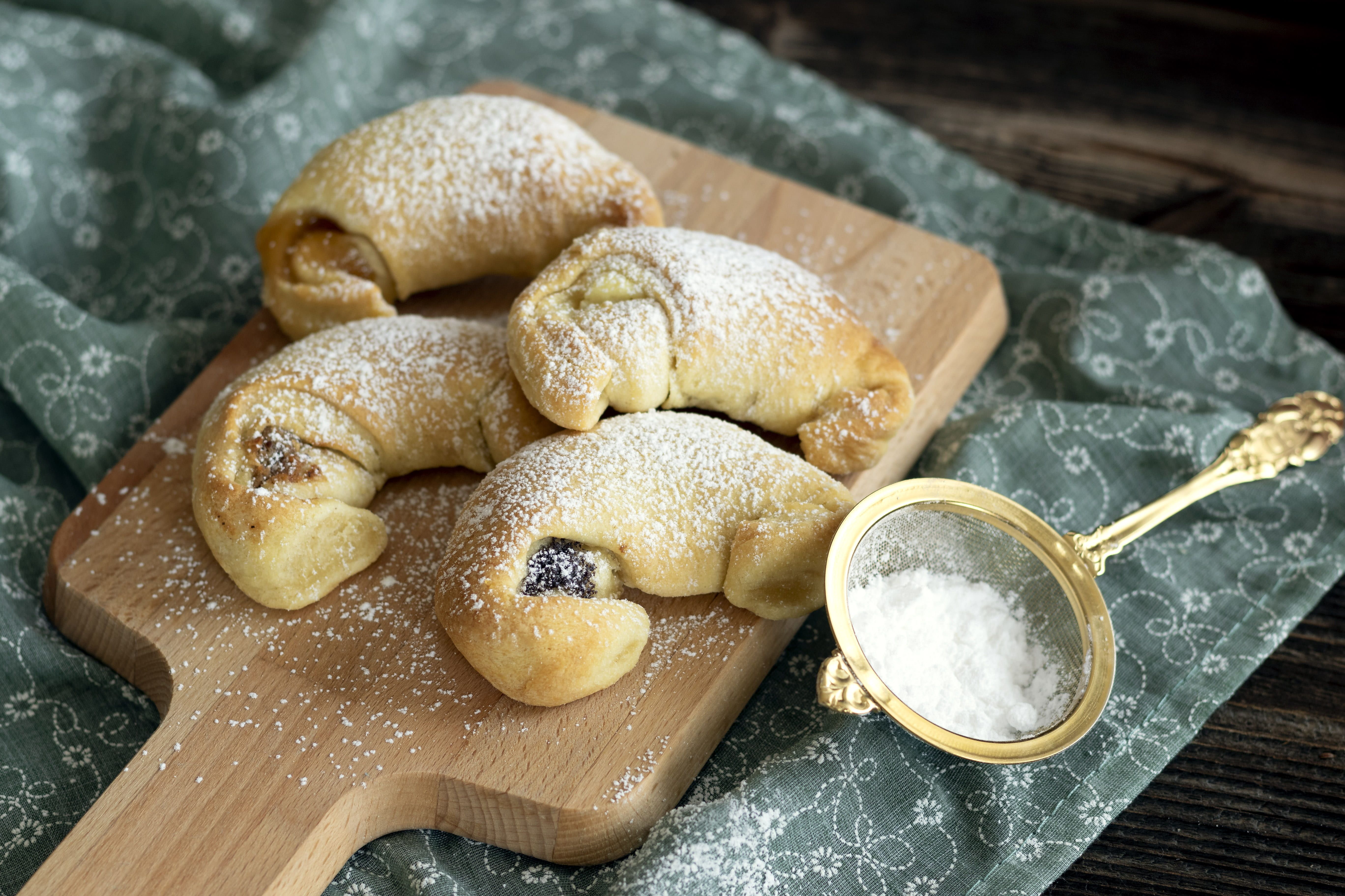 Négy krémes croissant porcukorral megszórt fatáblán, mellette porcukorral megszórt szitán.