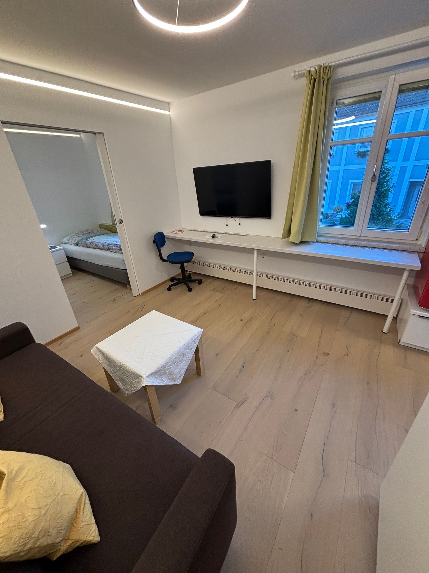 Modern nappali barna kanapéval, kis asztallal, TV-vel a falon, íróasztallal és székkel. Ablakából kilátás nyílik egy kék épületre.