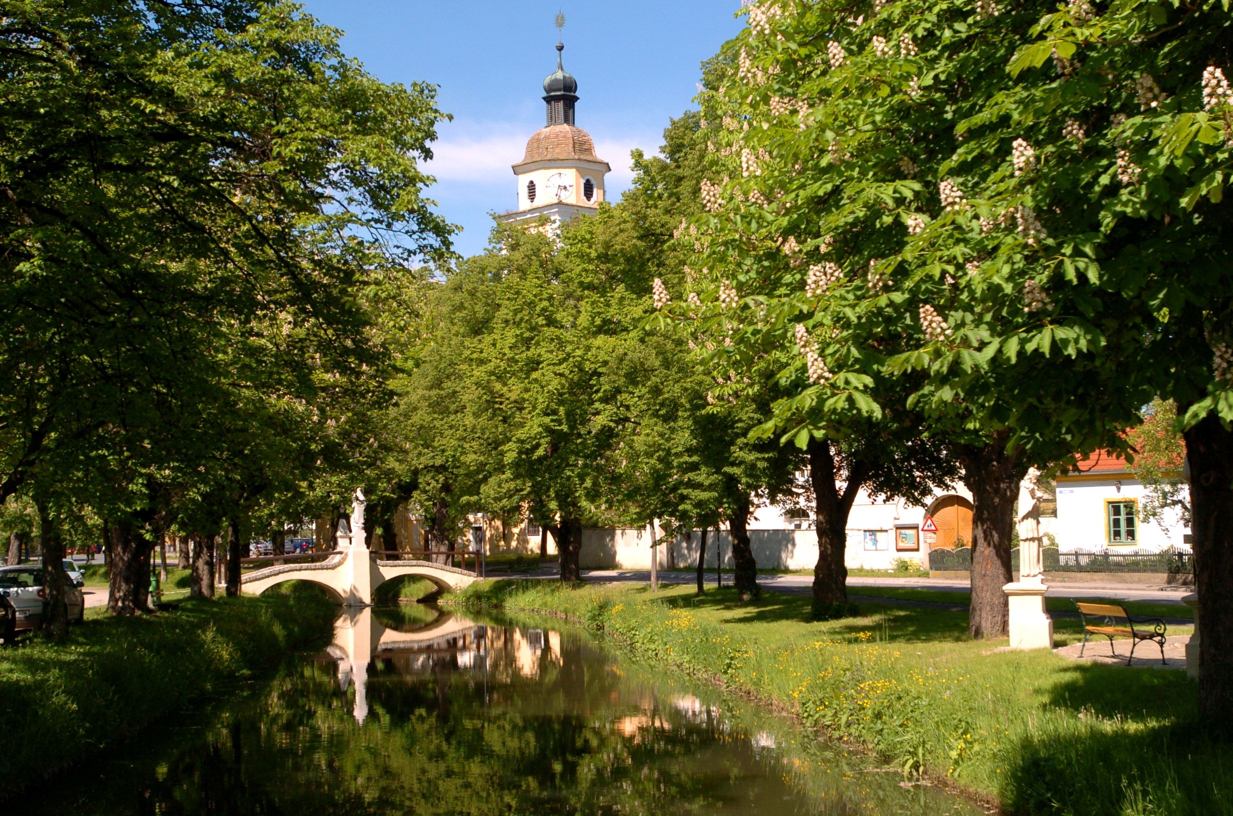 Idilli park, a háttérben egy kis folyó, egy híd és egy templomtorony.