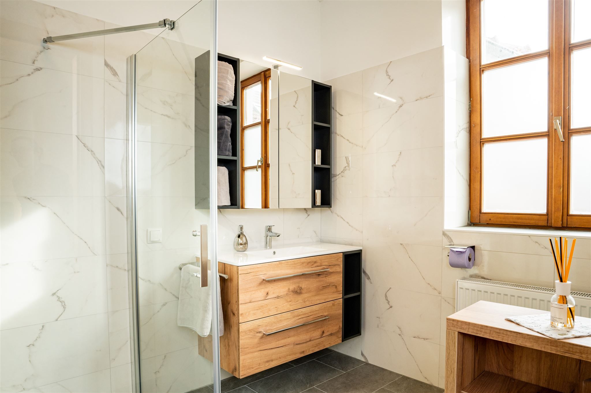 Modern fürdőszoba fabútorokkal, nagy ablakfronttal és üvegzuhannyal.