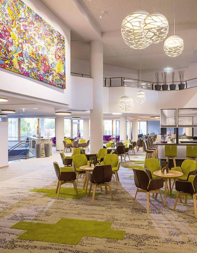Modern szálloda lobby szőnyegpadlóval, kerek asztalokkal, zöld székekkel, nagy lámpákkal és egy freskóval.