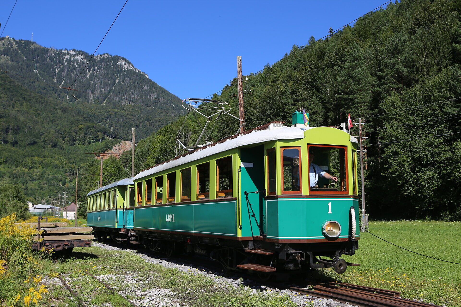 Schmalspurtriebwagen fährt auf einem Gleis durch eine sonnige Alpenlandschaft mit bewaldeten Hängen.