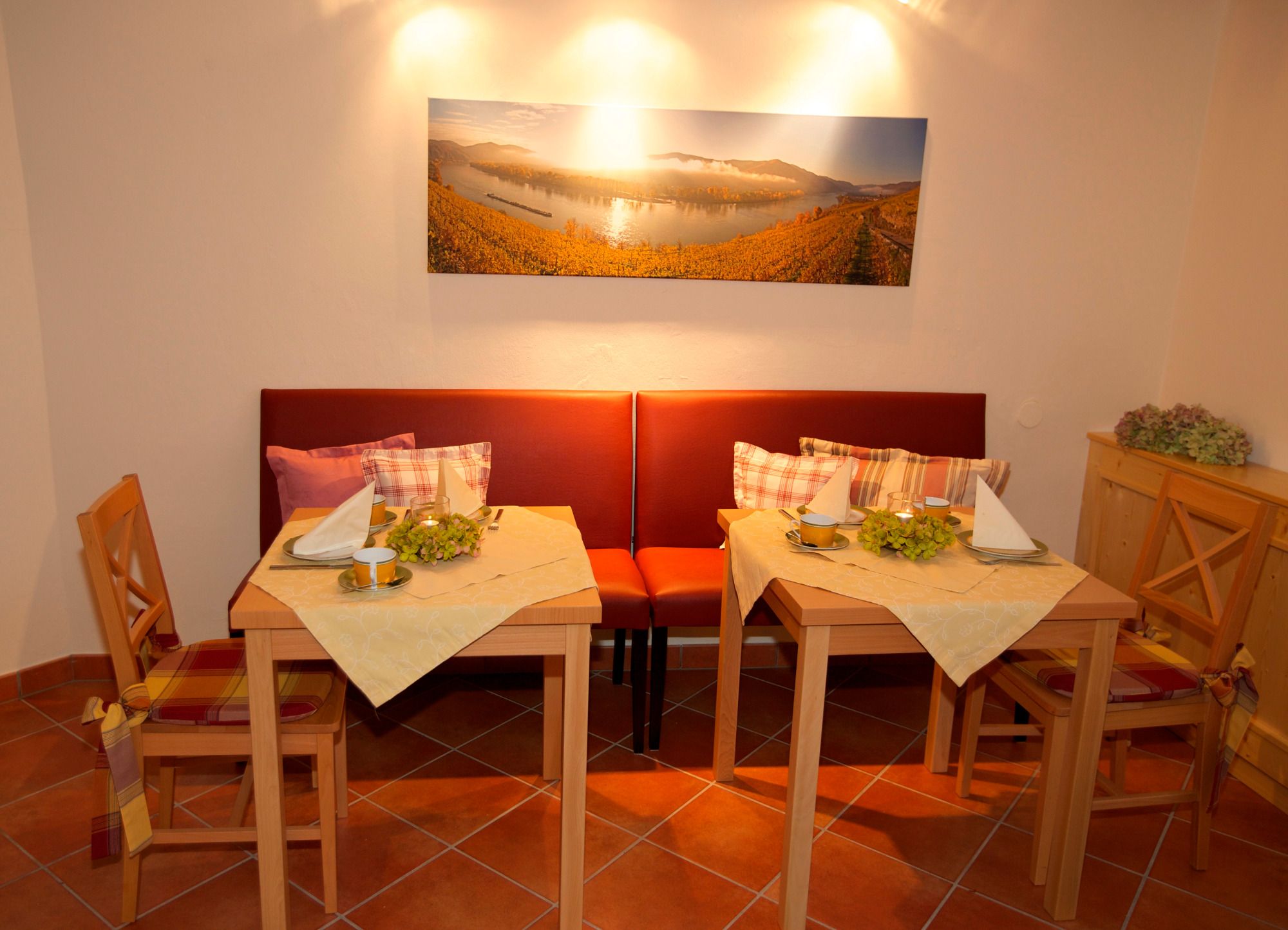 Hangulatos reggelizőszoba két terített asztallal, piros padokkal és egy tájképfestménnyel a falon.