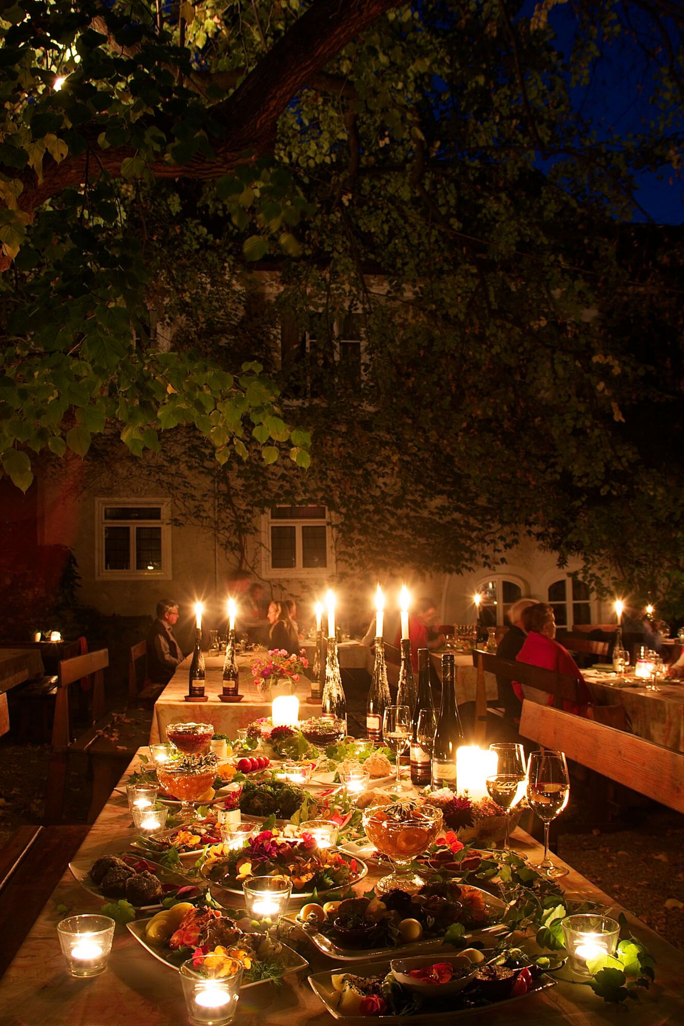 Ünnepélyesen megterített asztal a szabadban, gyertyafénnyel és különböző ételekből álló büféasztal. A háttérben emberek ülnek az asztaloknál.