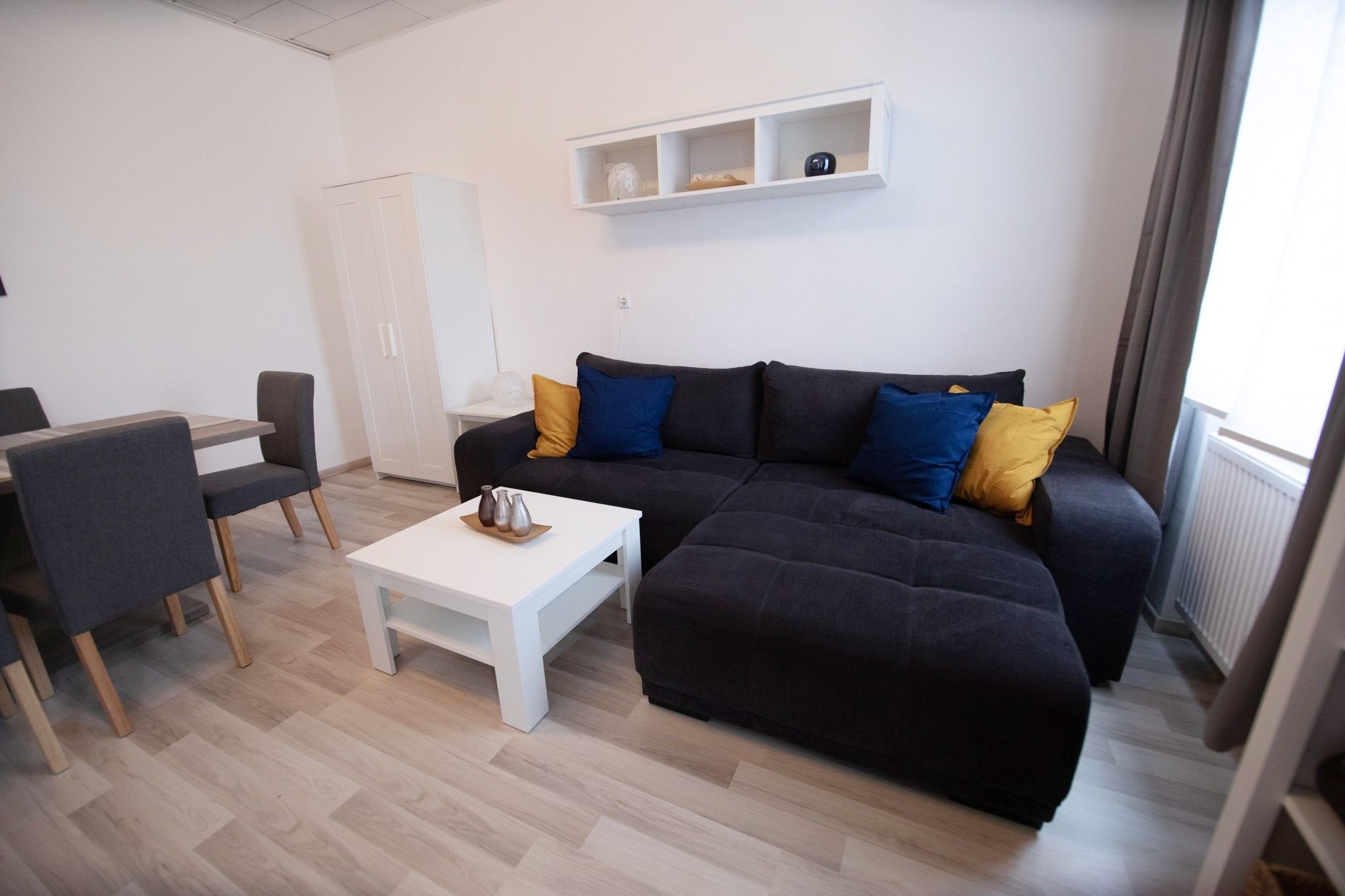 Modern nappali szürke kanapéval, színes párnákkal, fehér dohányzóasztallal és étkezőasztallal székekkel.