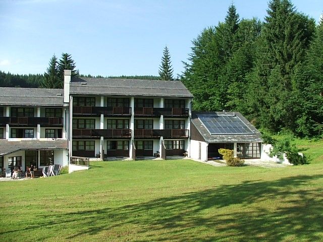 Hotelépület napelemekkel és zöld rét az előtérben, erdővel körülvéve.