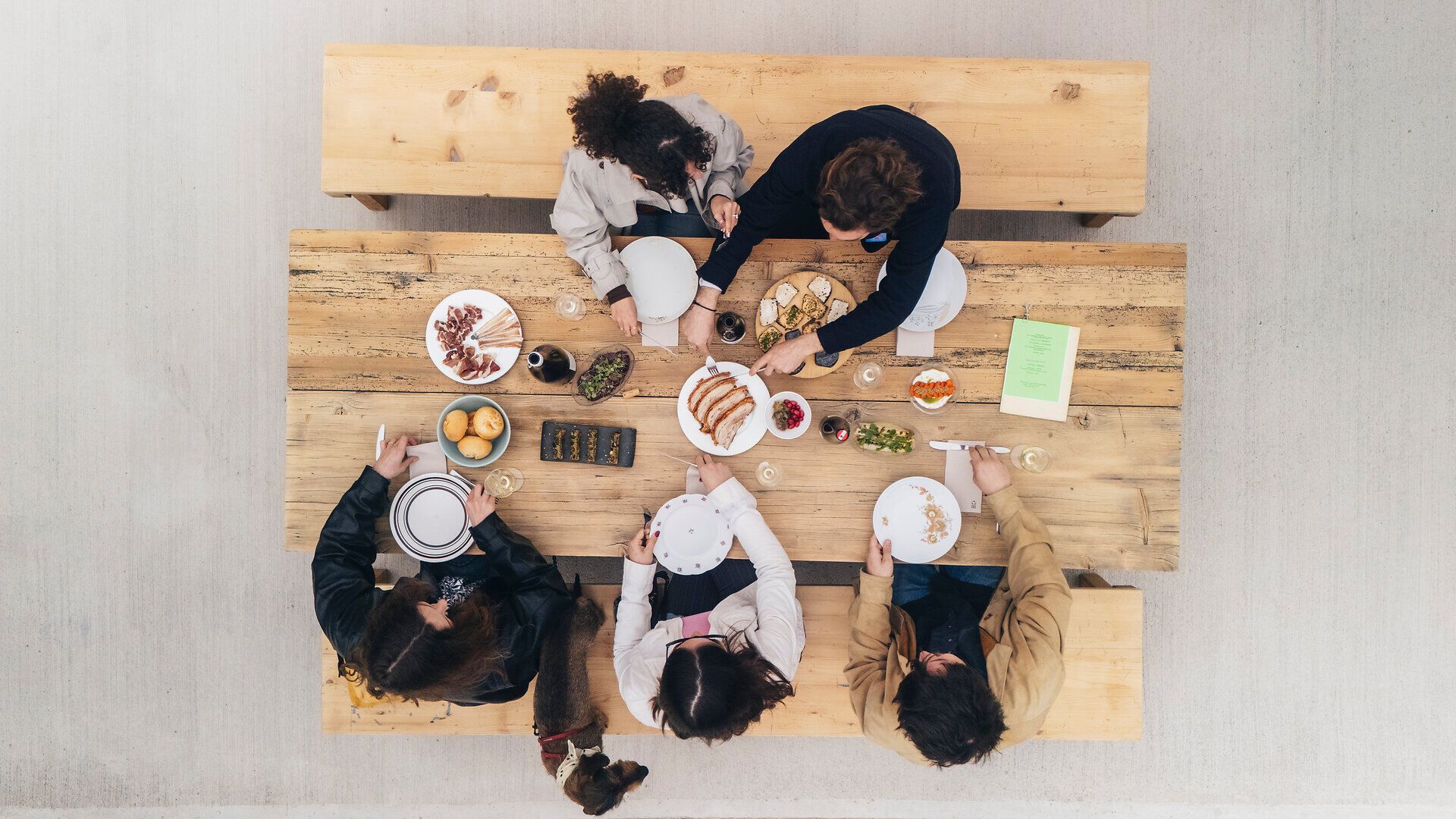 Draufsicht auf mehrere Personen, die an einem langen Holztisch sitzen; auf dem Tisch stehen Teller, Gläser und kleine Speisen, typisch für einen Heurigen.