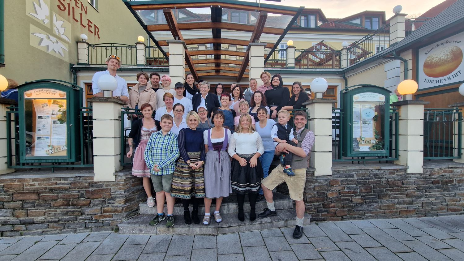 Emberek egy csoportja áll egy "Kirchenwirt" feliratú épület előtt.