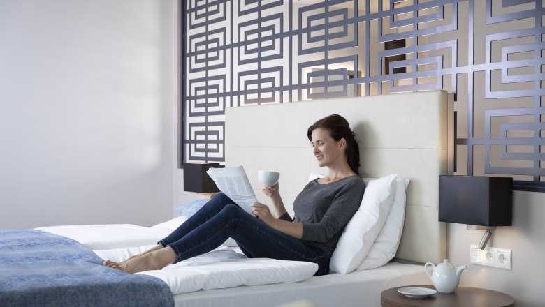 Nő pihen egy &aacute;gyon egy modern sz&aacute;llodai szob&aacute;ban, &uacute;js&aacute;got olvas &eacute;s egy cs&eacute;sz&eacute;t tart a kez&eacute;ben. A h&aacute;tt&eacute;rben dekorat&iacute;v fal.