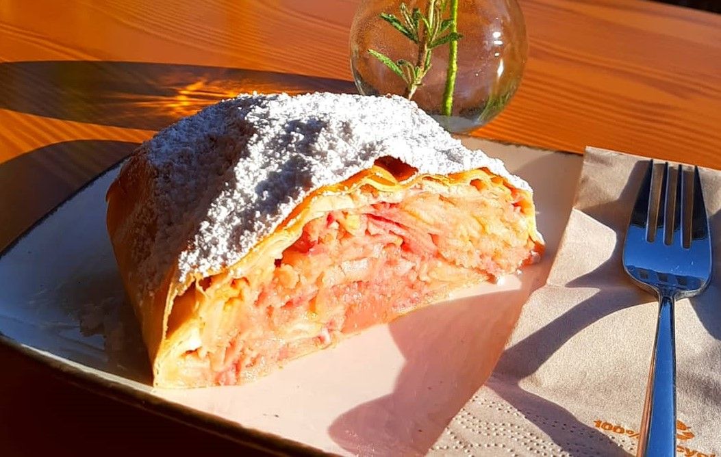 Egy szelet almás rétes porcukorral a tányéron, mellette egy villa és egy váza rozmaringgal.