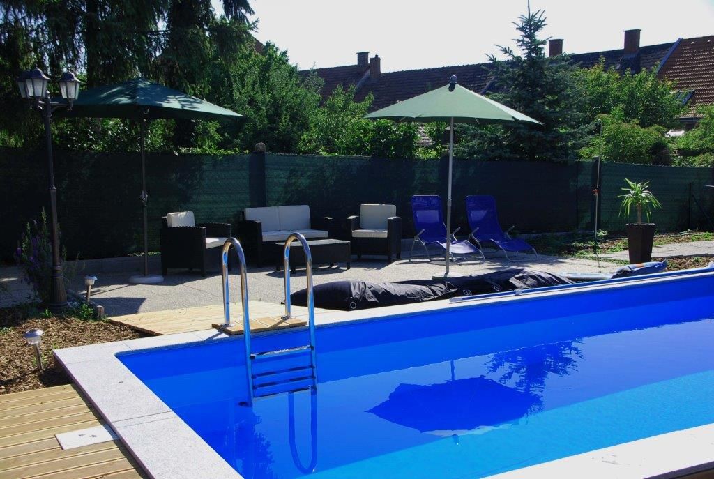 Kültéri terület kék medencével, amelyet napozóágyak, napernyők és kerti bútorok vesznek körül.