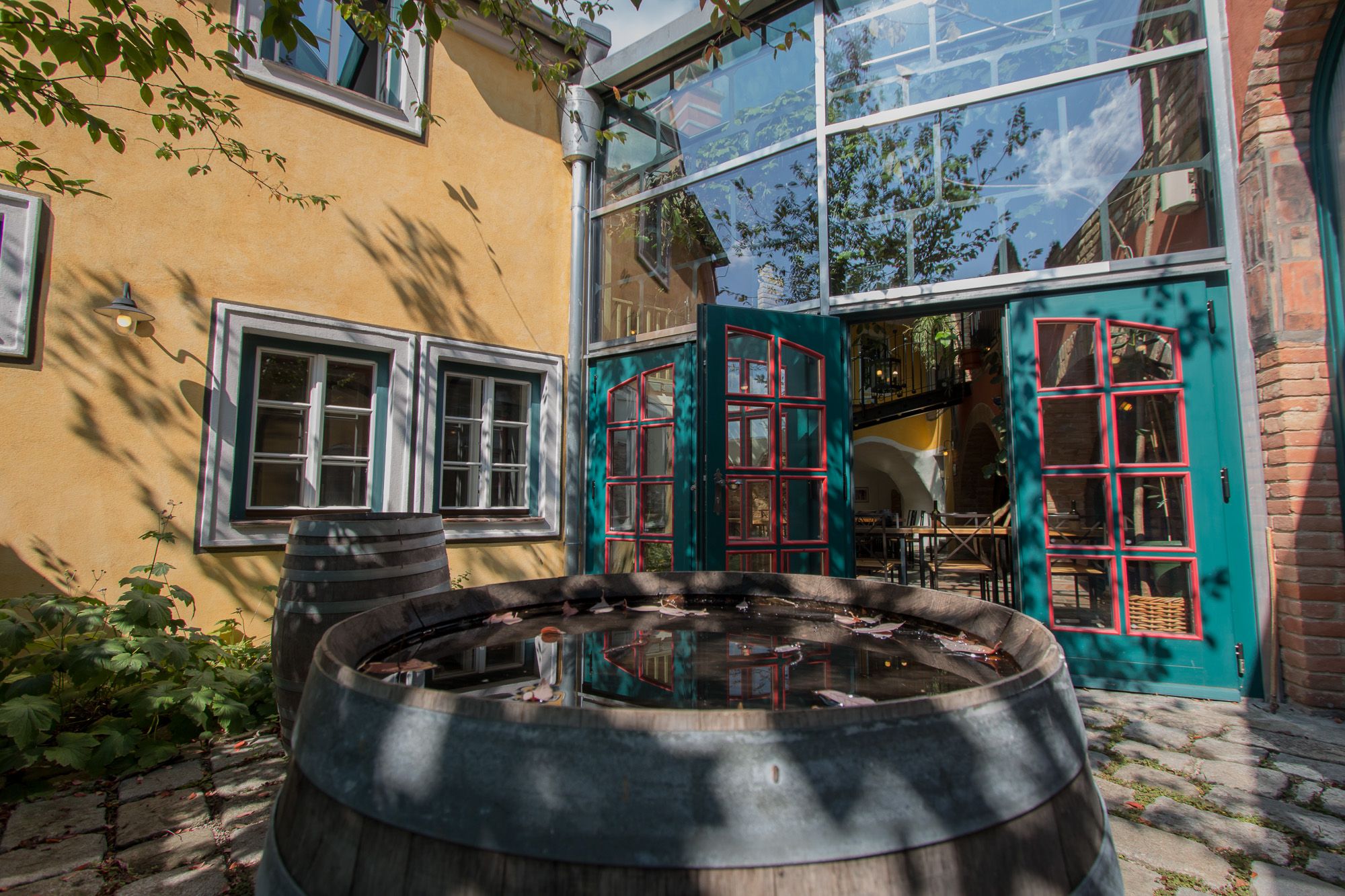 Egy belső udvar sárga homlokzattal, zöld ajtókkal és egy fahordó vízzel. A falra fák árnyai vetülnek.