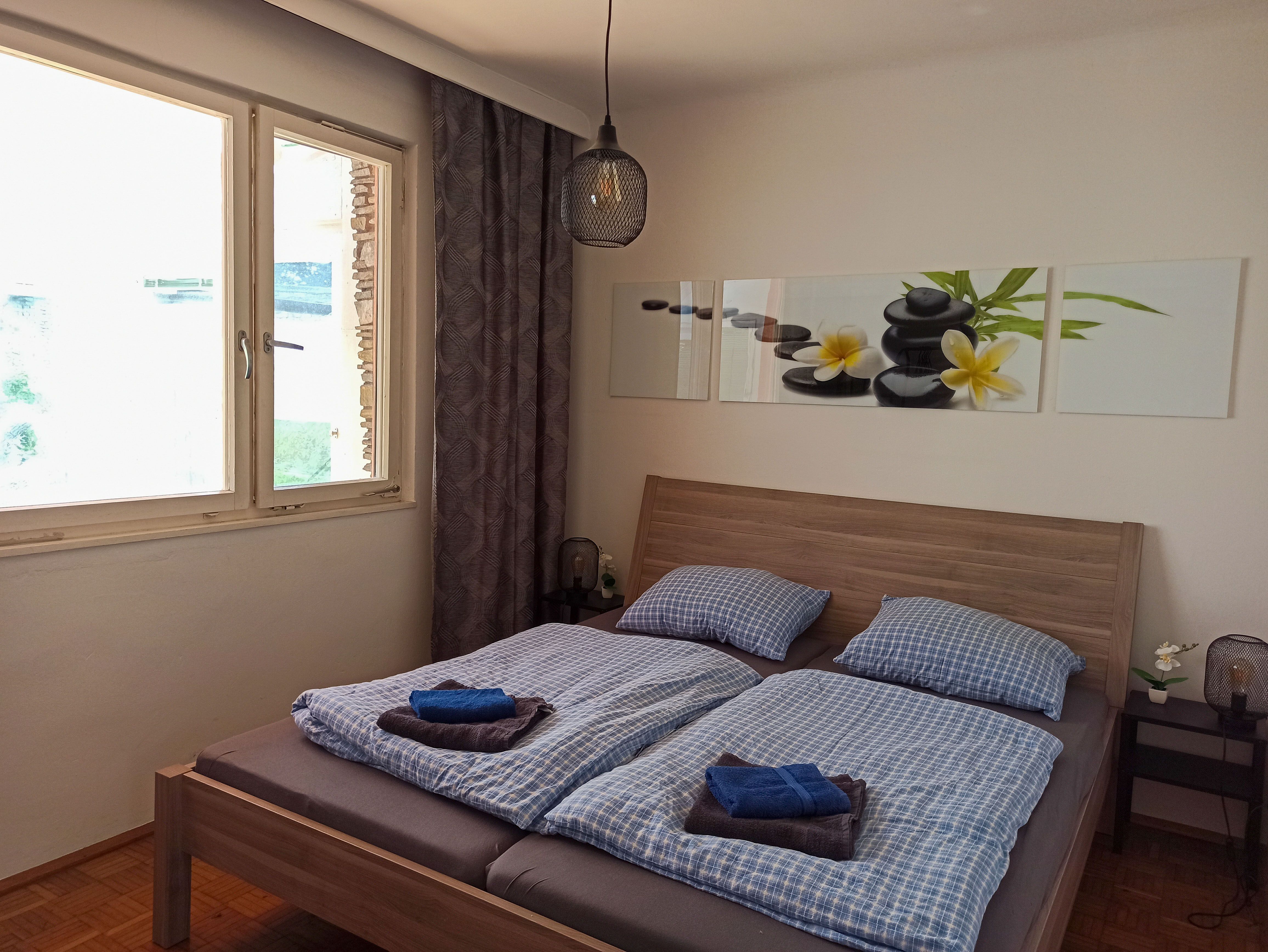 Hálószoba franciaággyal, kék ágyneművel, kő- és virágos képpel a falon, ablakkal és éjjeli lámpával.